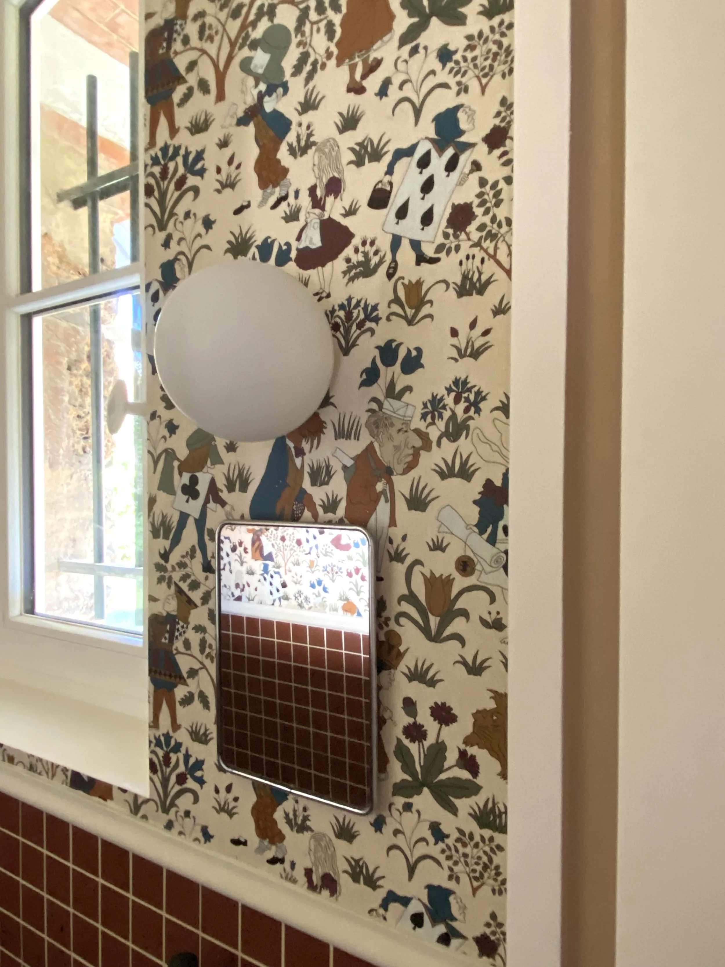 Détail d'un mur tapissé d'un papier peint House of Hackney représenatnt Alice au pays des merveilles, d'un miroir de barbier chiné, d'un luminaire boule en verre et de carrelage en soubassement à petits carreaux rouges