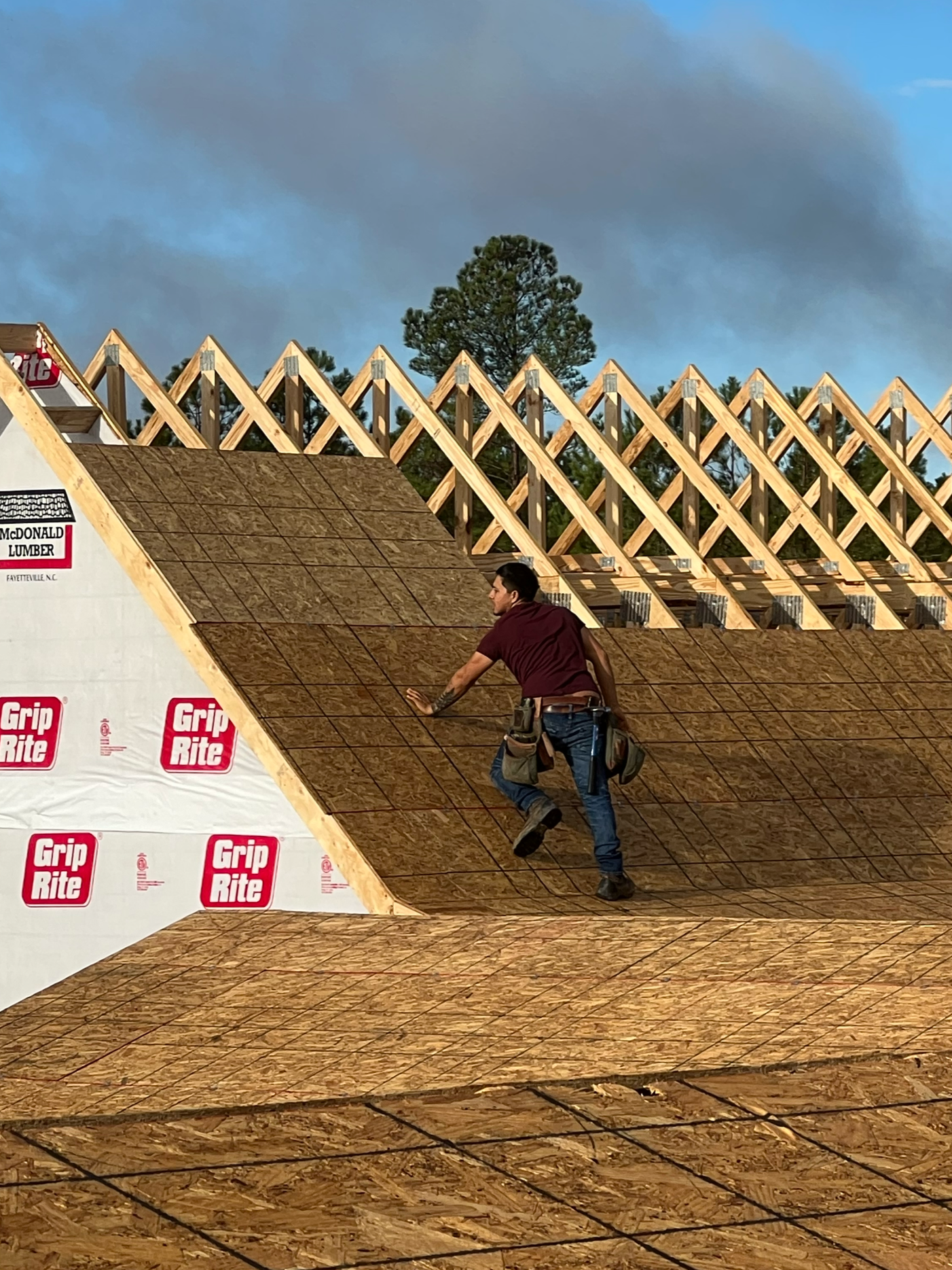 A construction worker installing roof sheathing on a building under construction with a partly cloudy sky in the background.