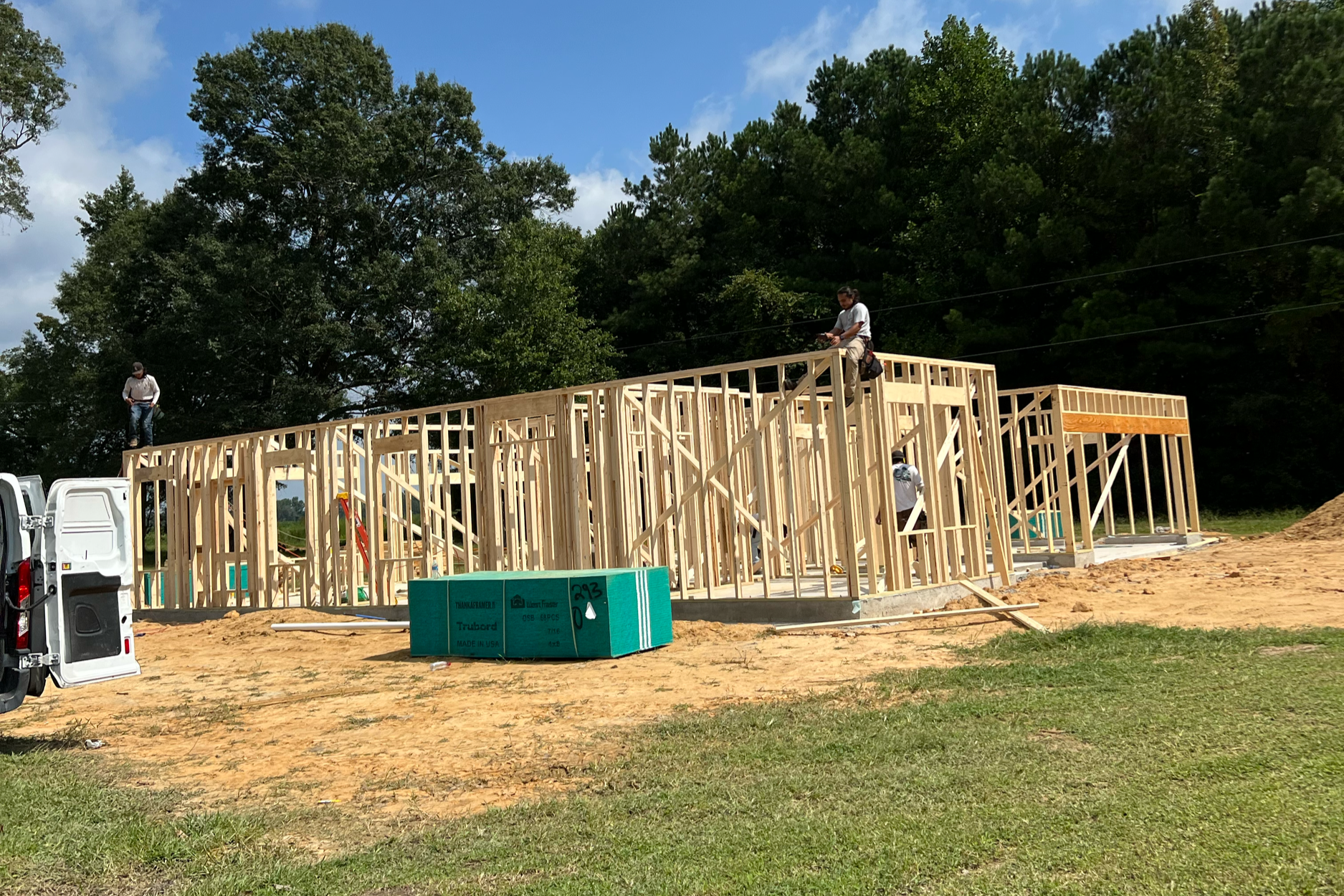 Construction workers building a house with wooden framing on a construction site surrounded by trees and a blue sky.