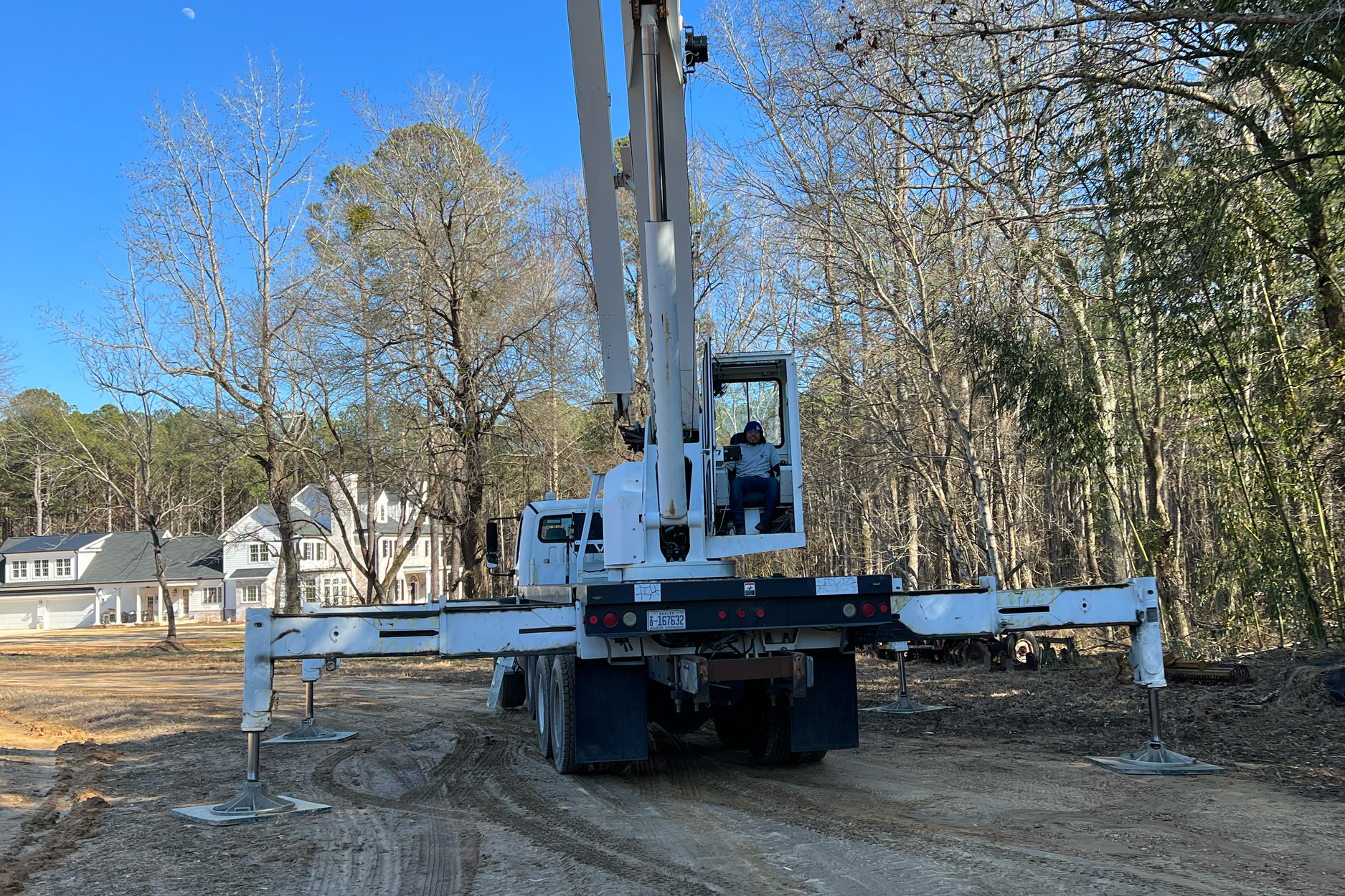 A white crane truck with extended arm and stabilizers is on a construction site with dirt ground, surrounded by leafless trees and residential houses in the background, under a clear blue sky.
