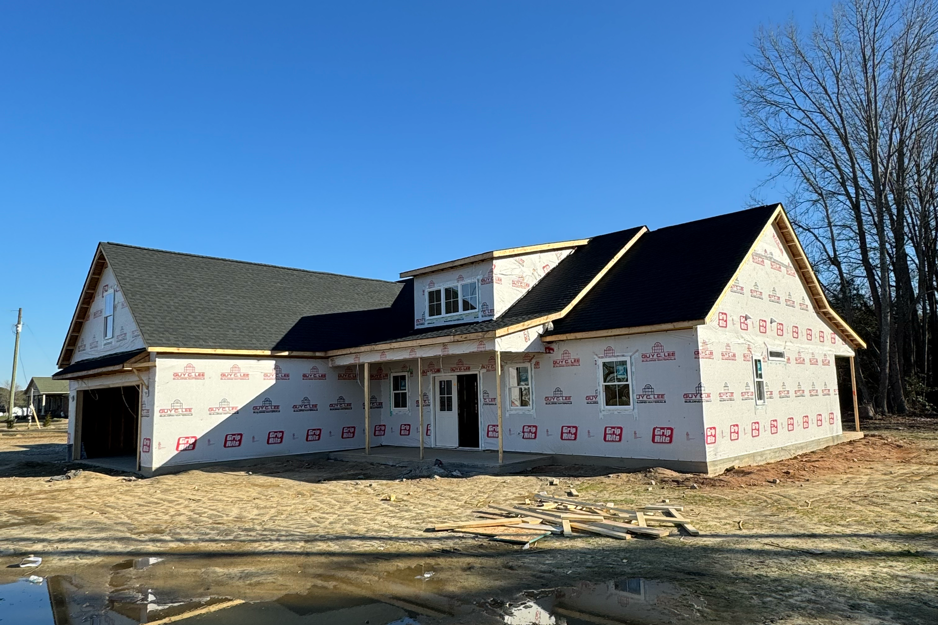 Construction house with partially installed roofing and wrapped exterior walls, surrounded by dirt and a small pool of water, under a clear blue sky with bare trees in the background.