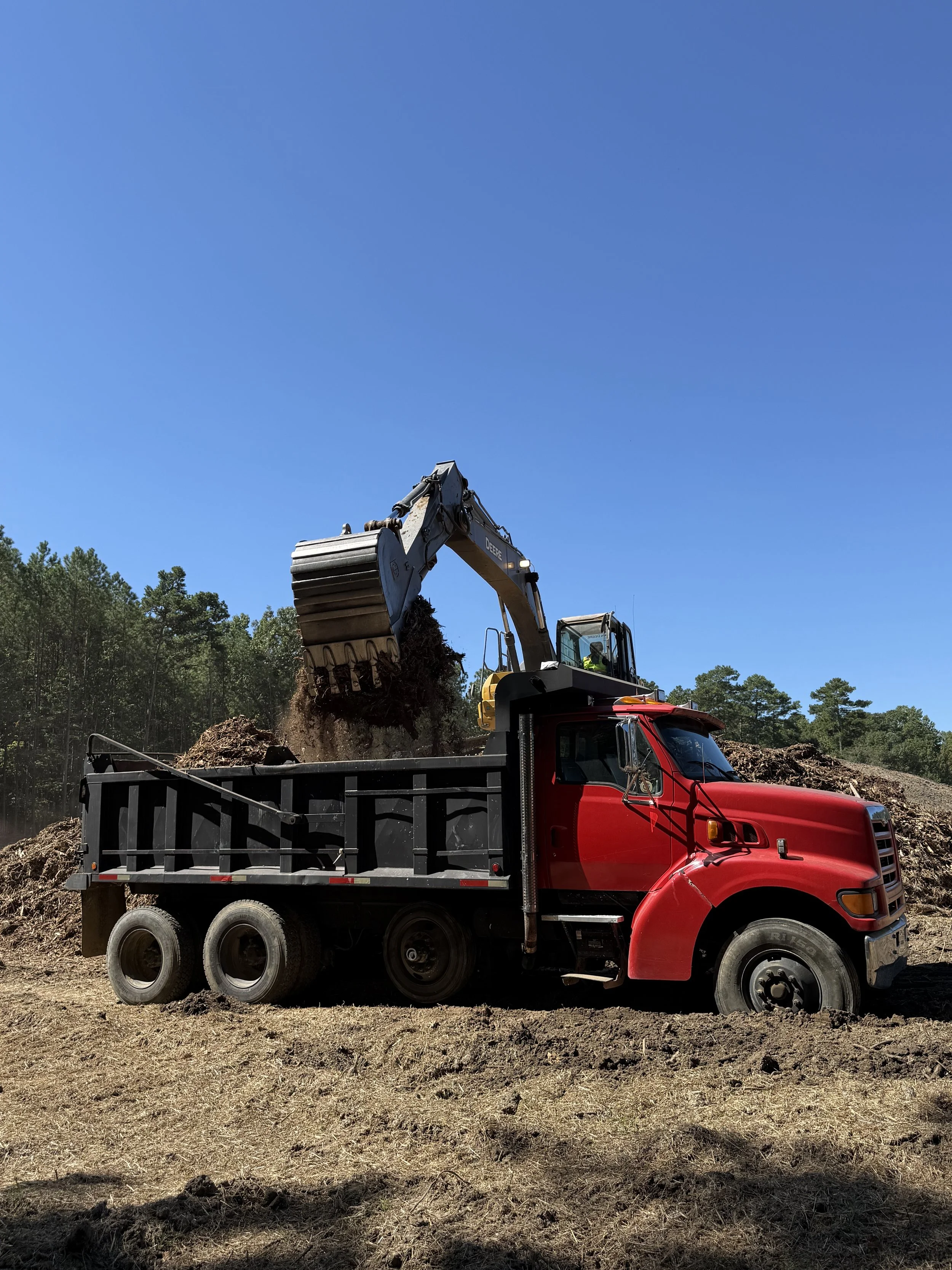 A red dump truck being loaded with soil or dirt by a large excavator at a construction site on a clear, sunny day, with trees visible in the background.