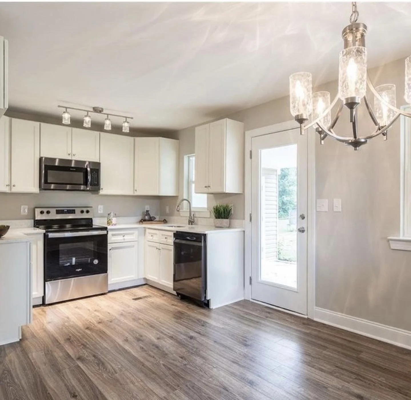 Bright kitchen with white cabinets, stainless steel appliances, Wood flooring, small window above sink, glass door leading outside, modern chandelier