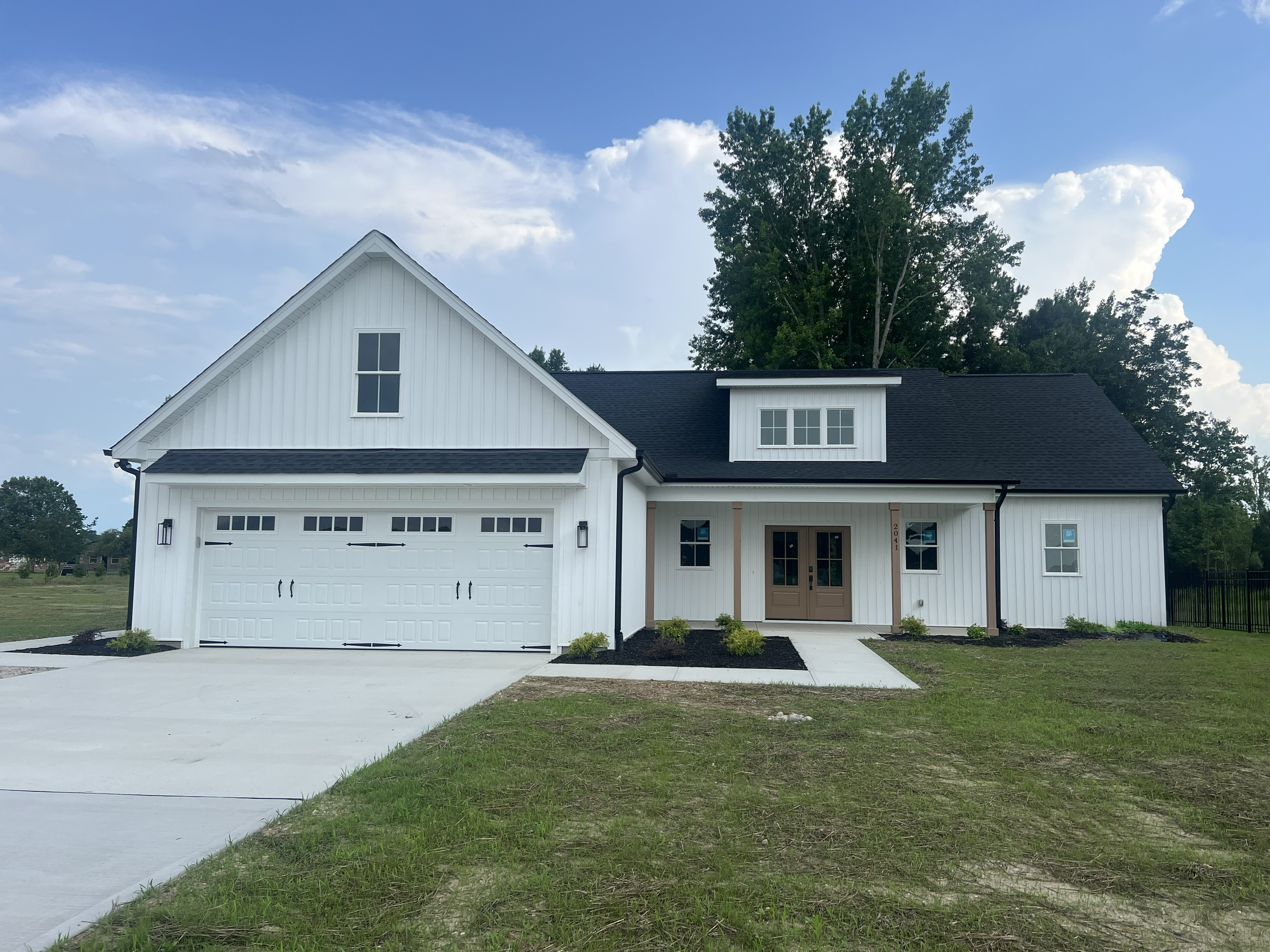 Modern white house with black roof, attached garage, front porch with brown double doors, and a small garden with shrubs, set against a background of trees and sky.