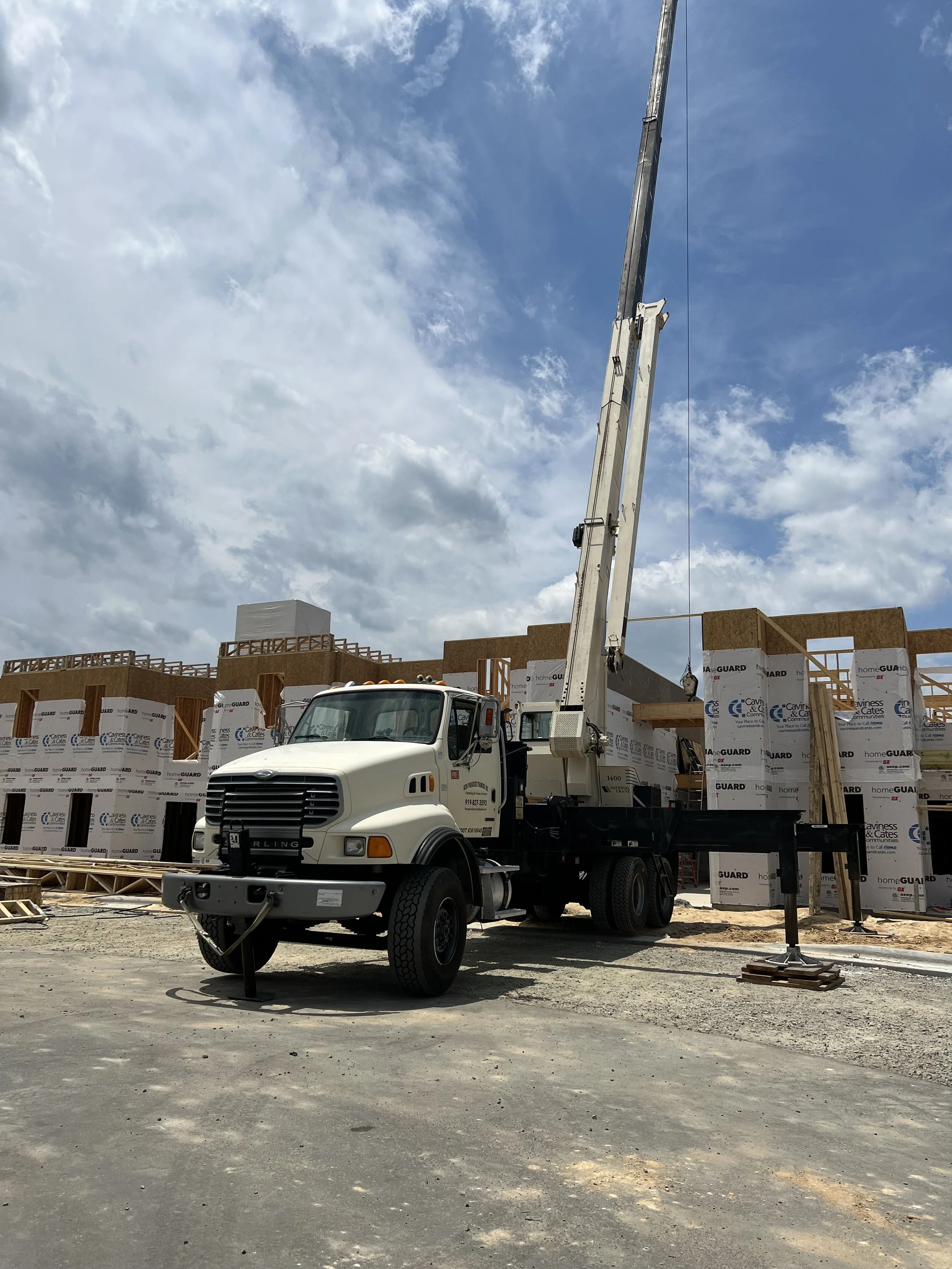 Construction site with a crane truck lifting materials in front of a partially constructed building with wood framing and insulation panels.