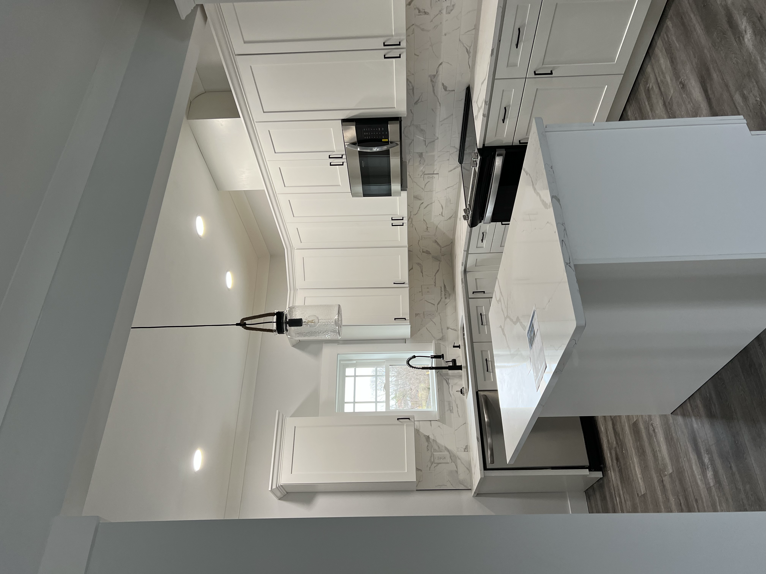 Modern white kitchen with marble countertops, black hardware, a built-in microwave, a black stove, a black faucet, and a large island with a marble top. Recessed lighting and a small window provide natural and artificial light.