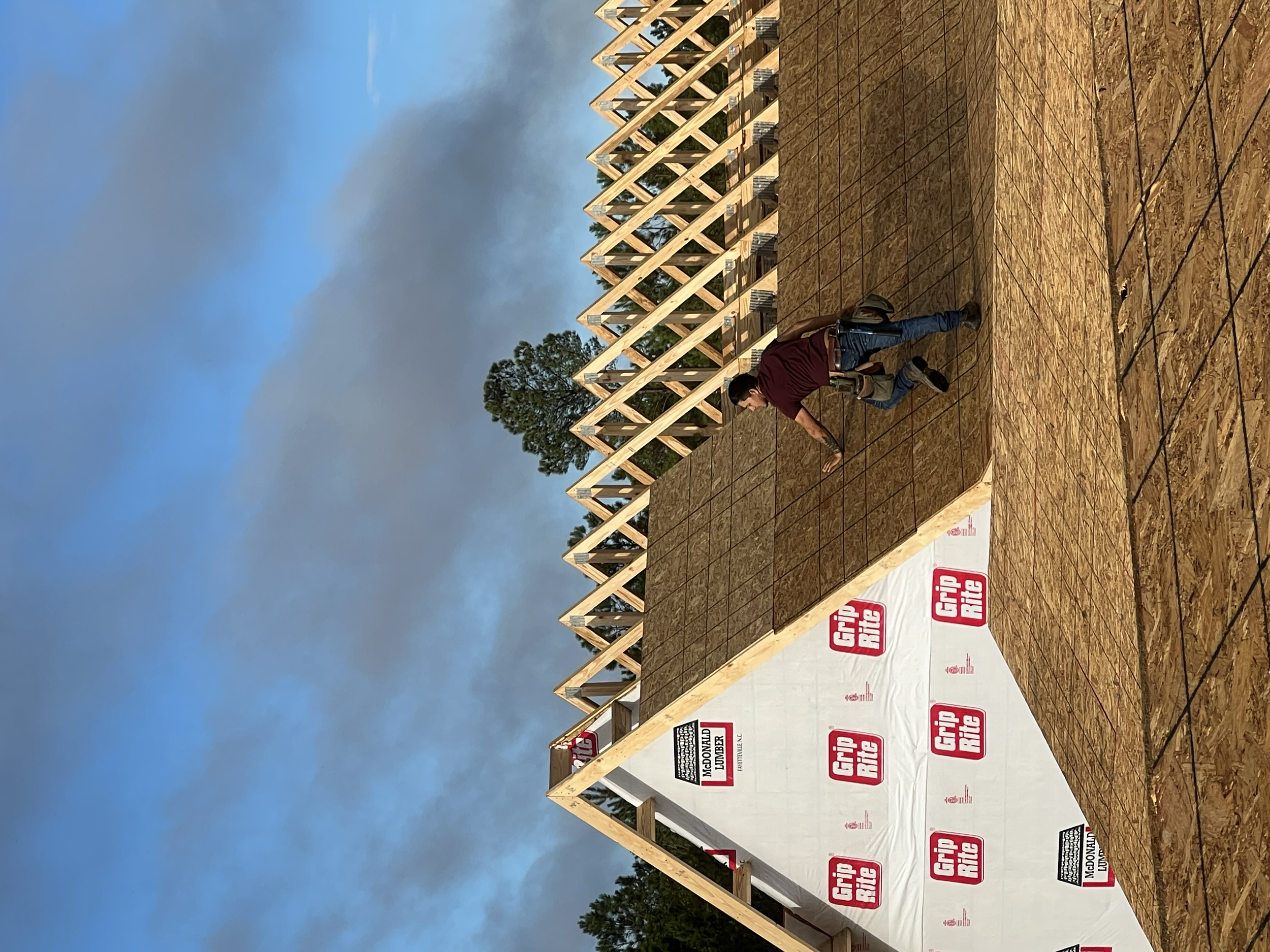 A construction worker is on a partially built house frame, installing plywood sheathing on the roof. The house has a wooden frame with both the roof and walls in progress. The area around the house shows a cloudy sky with patches of blue and some gre