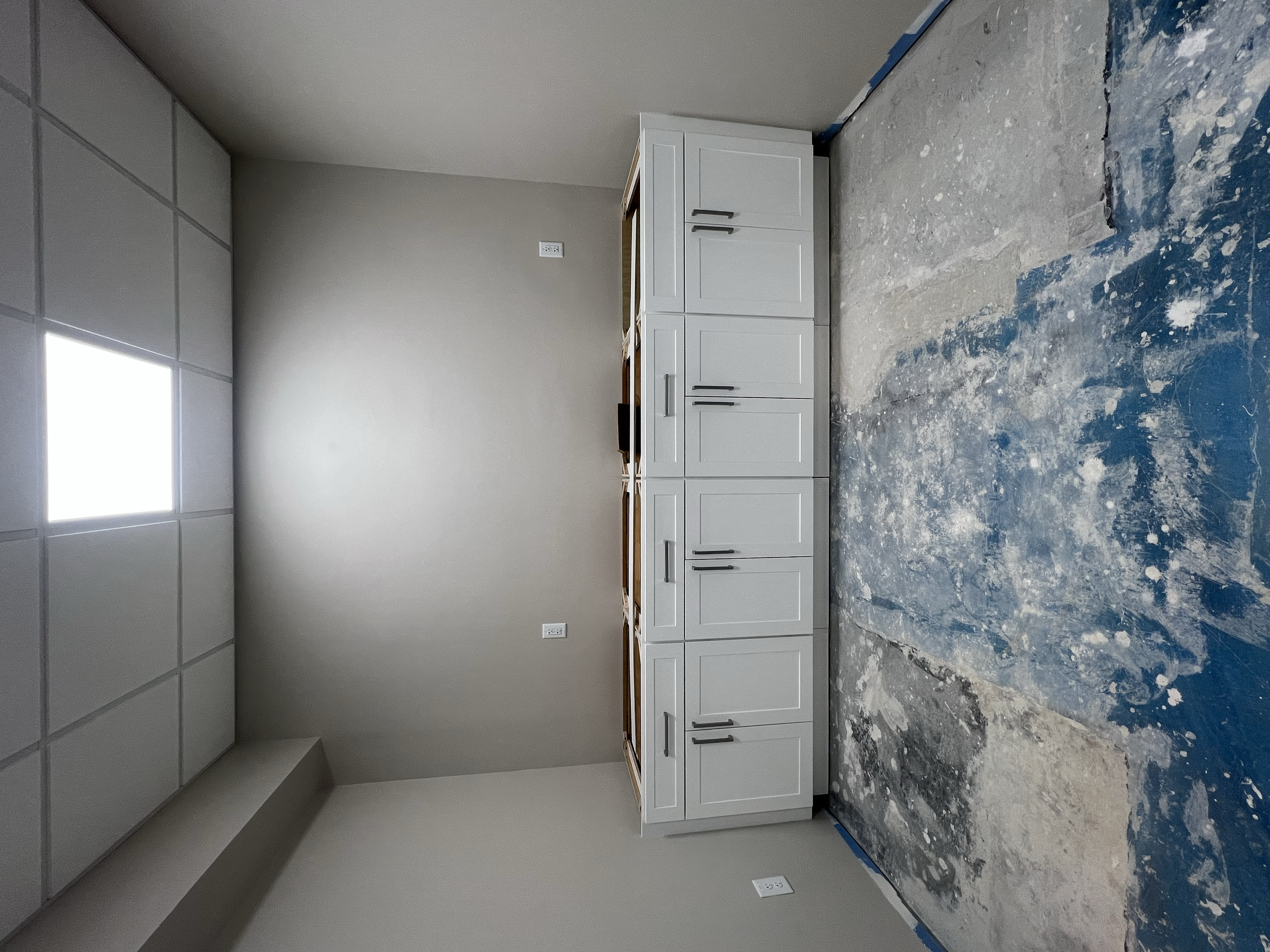 A kitchen under construction with white cabinets, a gray wall, and a concrete floor with paint splatters.
