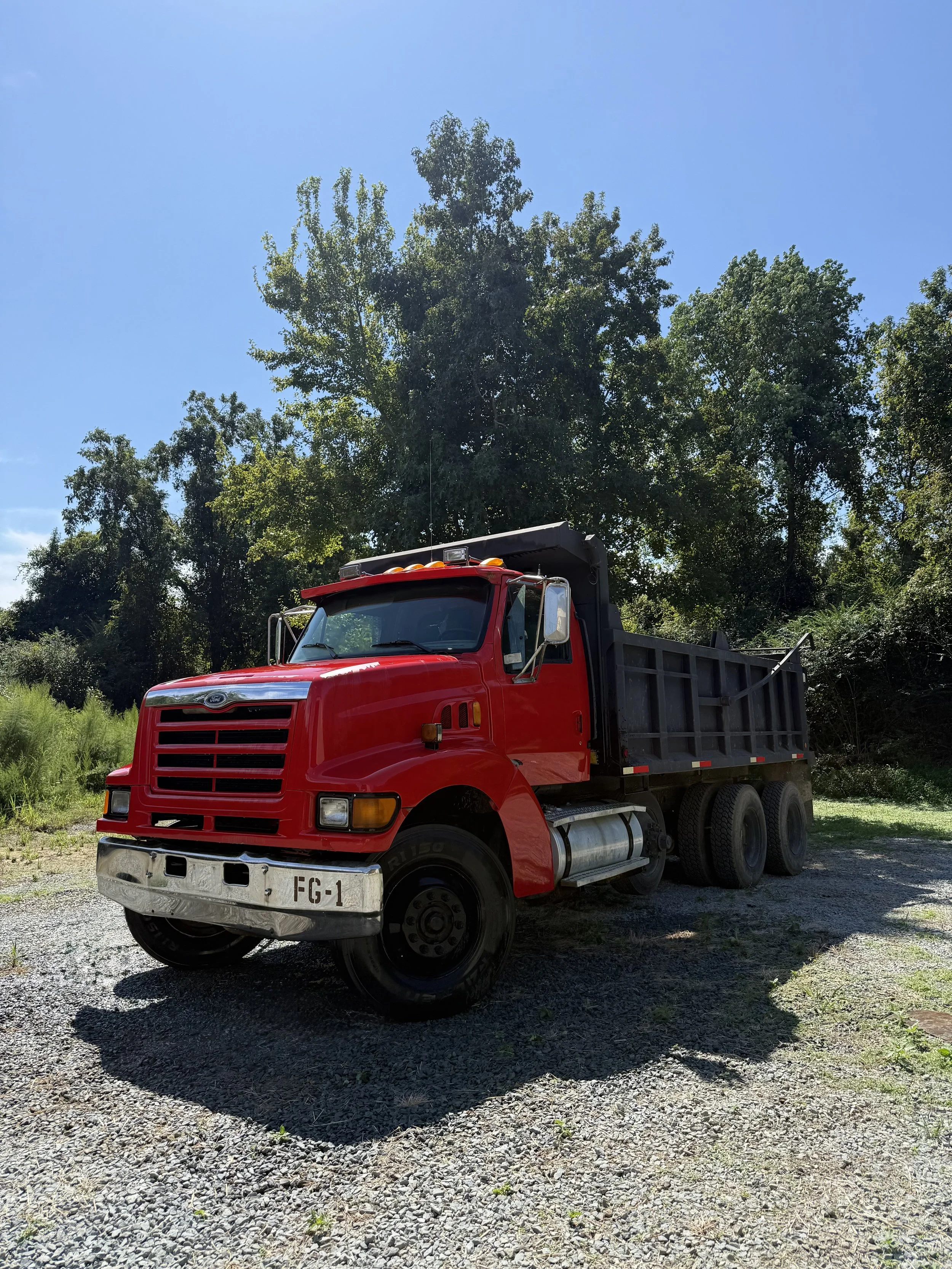 Red dump truck parked on gravel with trees and blue sky in the background.