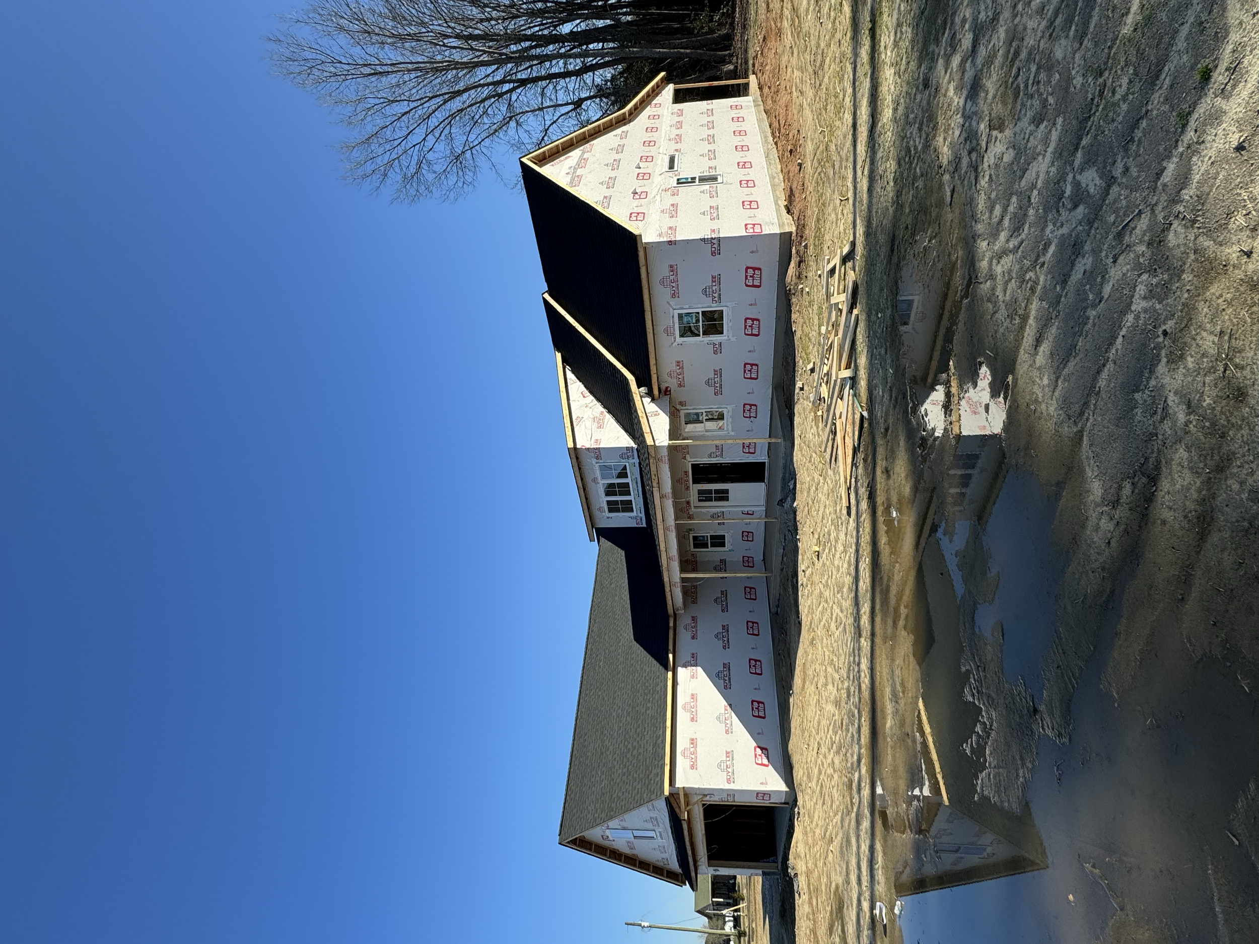 House under construction with unfinished exterior walls and a black roof, situated on a muddy area with puddles of water and construction materials scattered around, against a clear blue sky with a leafless tree in the background.