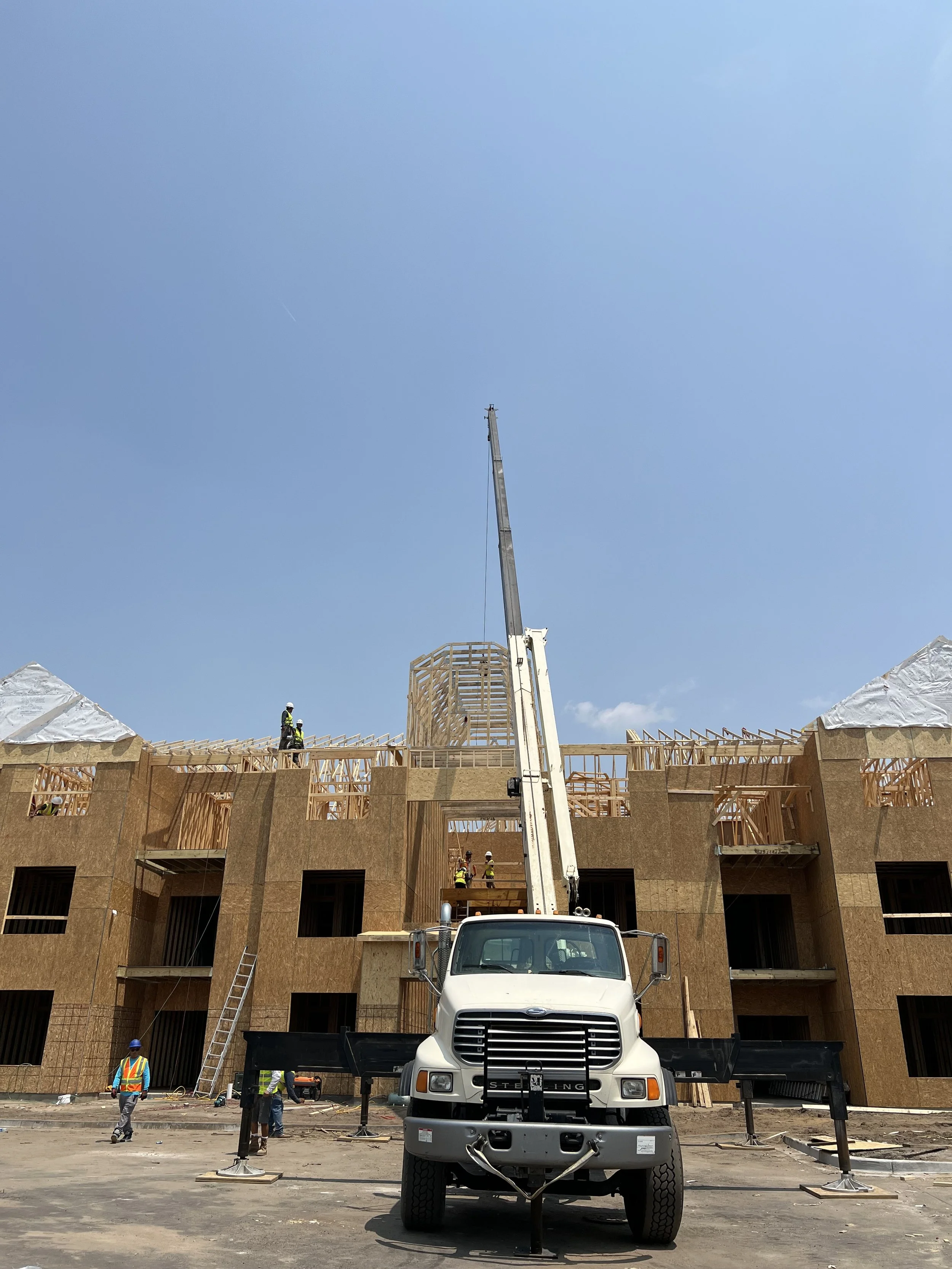 Construction site with workers building a wooden structure; a crane truck is centered in the foreground, lifting materials, with blue sky overhead.