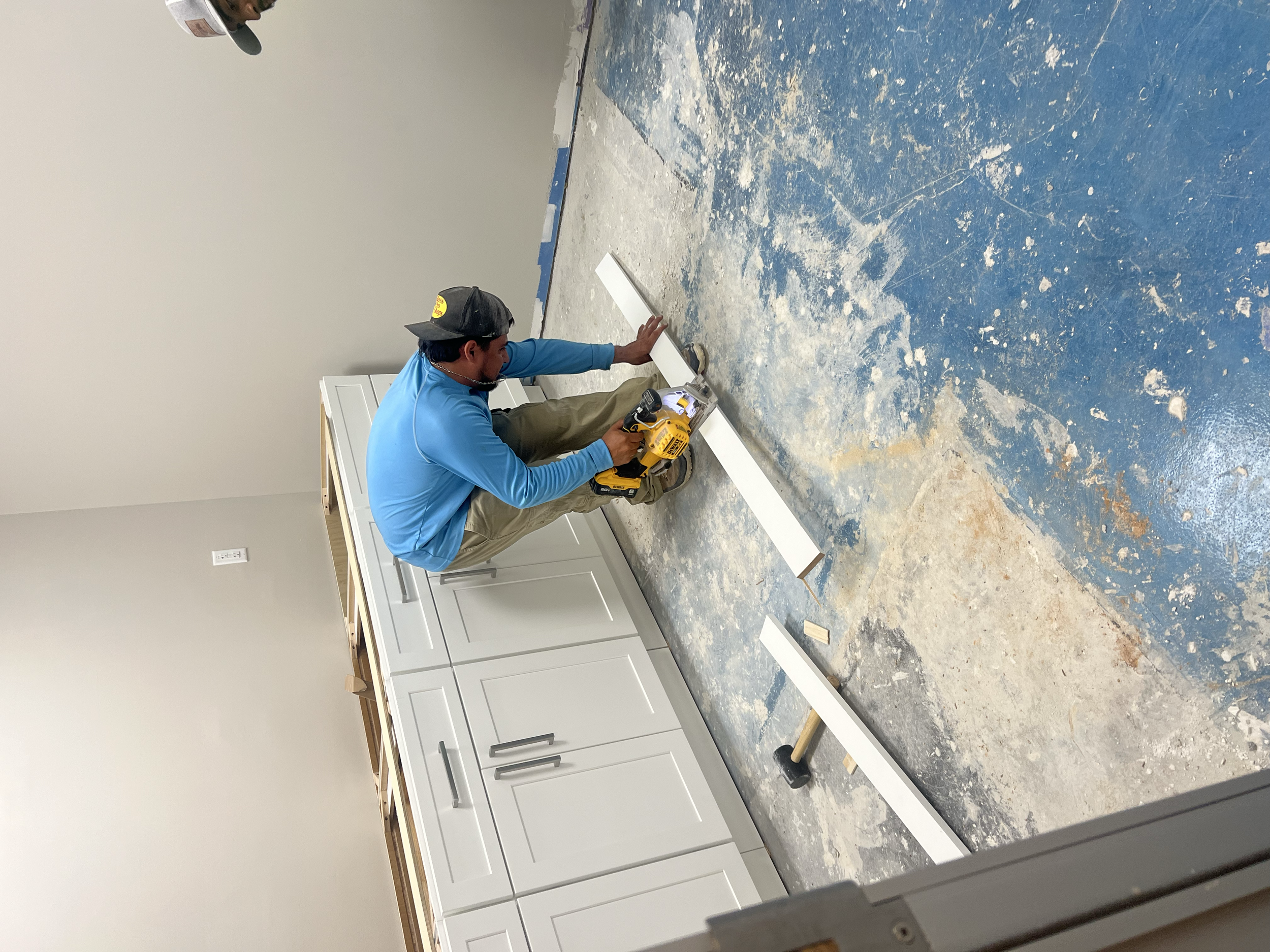A man installing a baseboard using a cordless saw in a room with unfinished flooring and white kitchen cabinets.