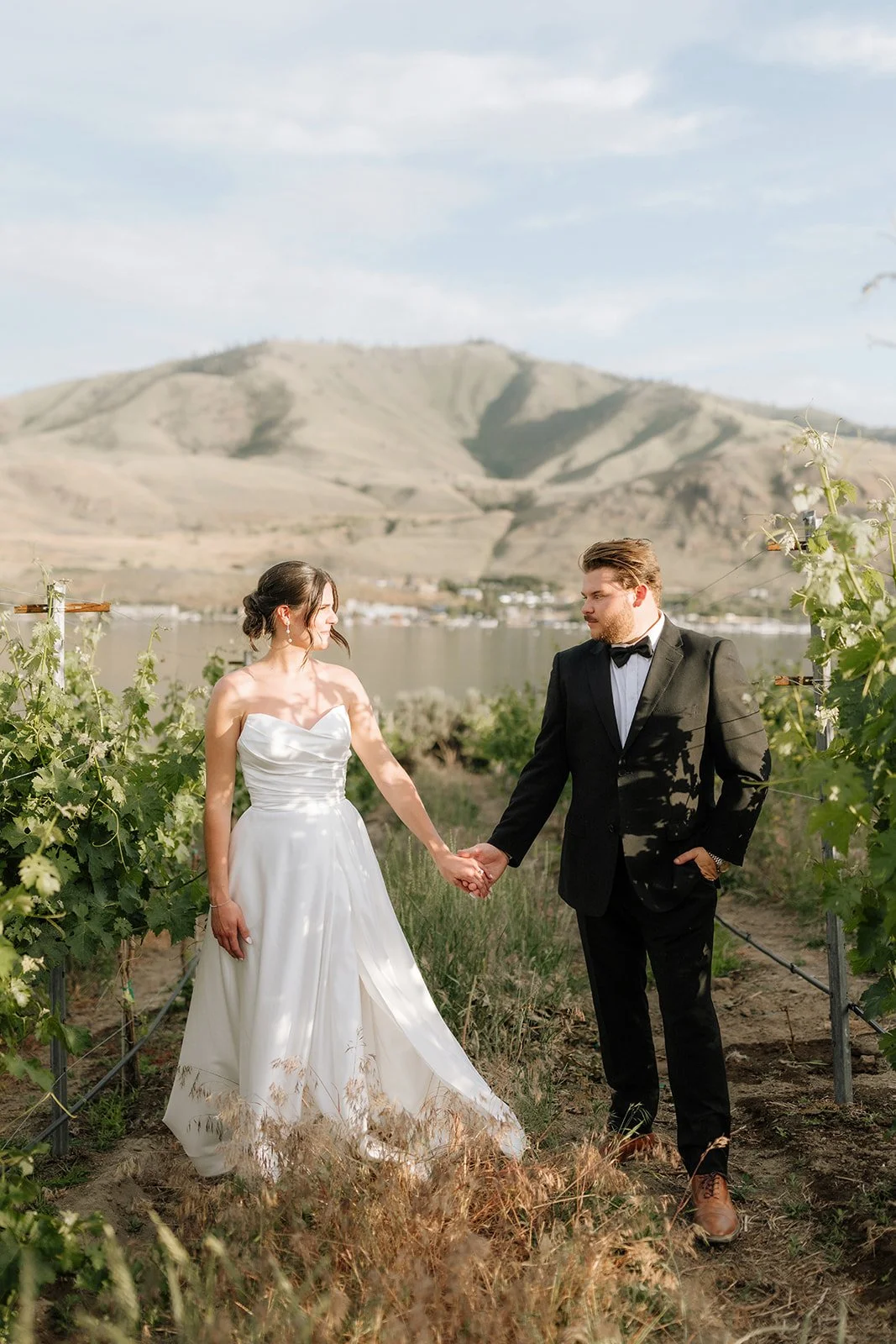 Bride and groom holding hands in a vineyard with mountains and a body of water in the background.