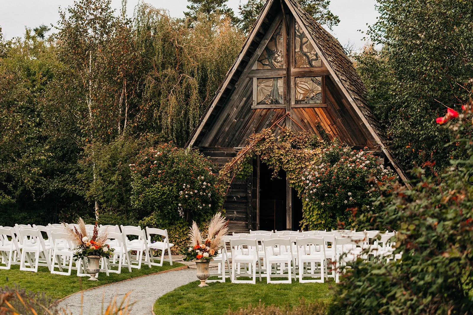 Outdoor wedding setup with white chairs and floral arrangements in urns, in front of a rustic wooden A-frame barn surrounded by lush trees and bushes.