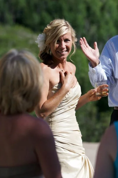 A woman in a white dress holding a drink, smiling, outdoors at a social gathering.
