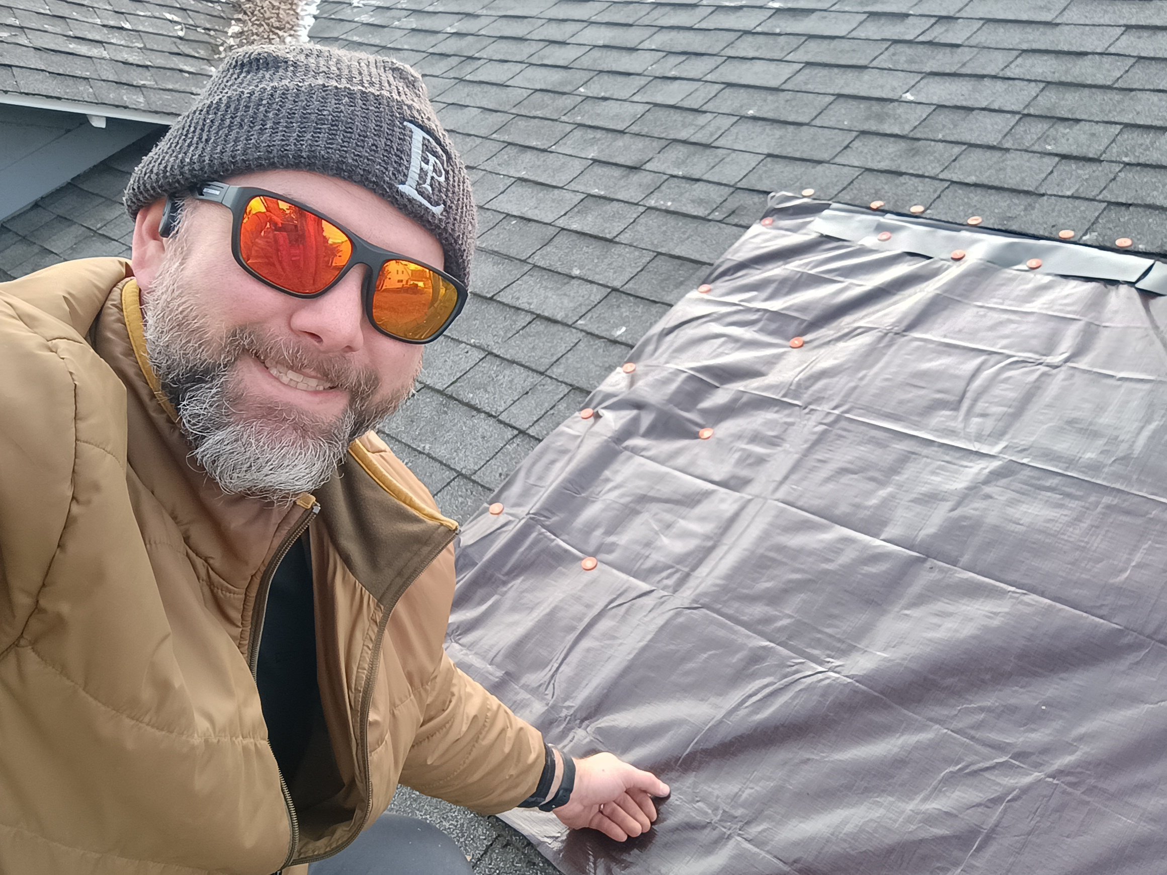 Owner, Steve Hutchinson, wearing a brown jacket, gray knit beanie, and sunglasses taking a selfie on a roof next to a large hole covered securely with copper-colored fasteners.