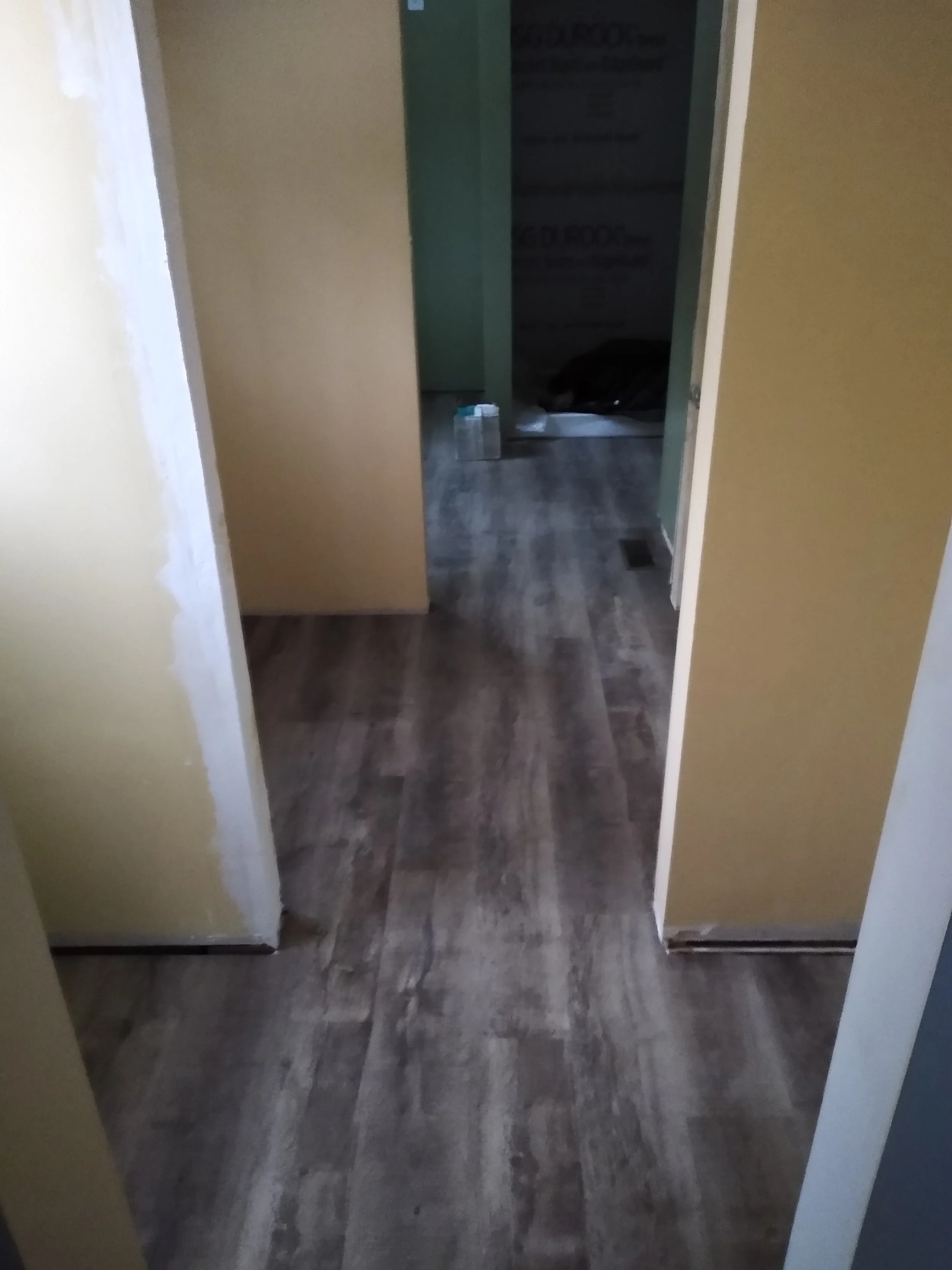Interior view of a house under renovation showing yellow and green walls, a partially visible doorway, and wood-look flooring with some construction supplies in the background.