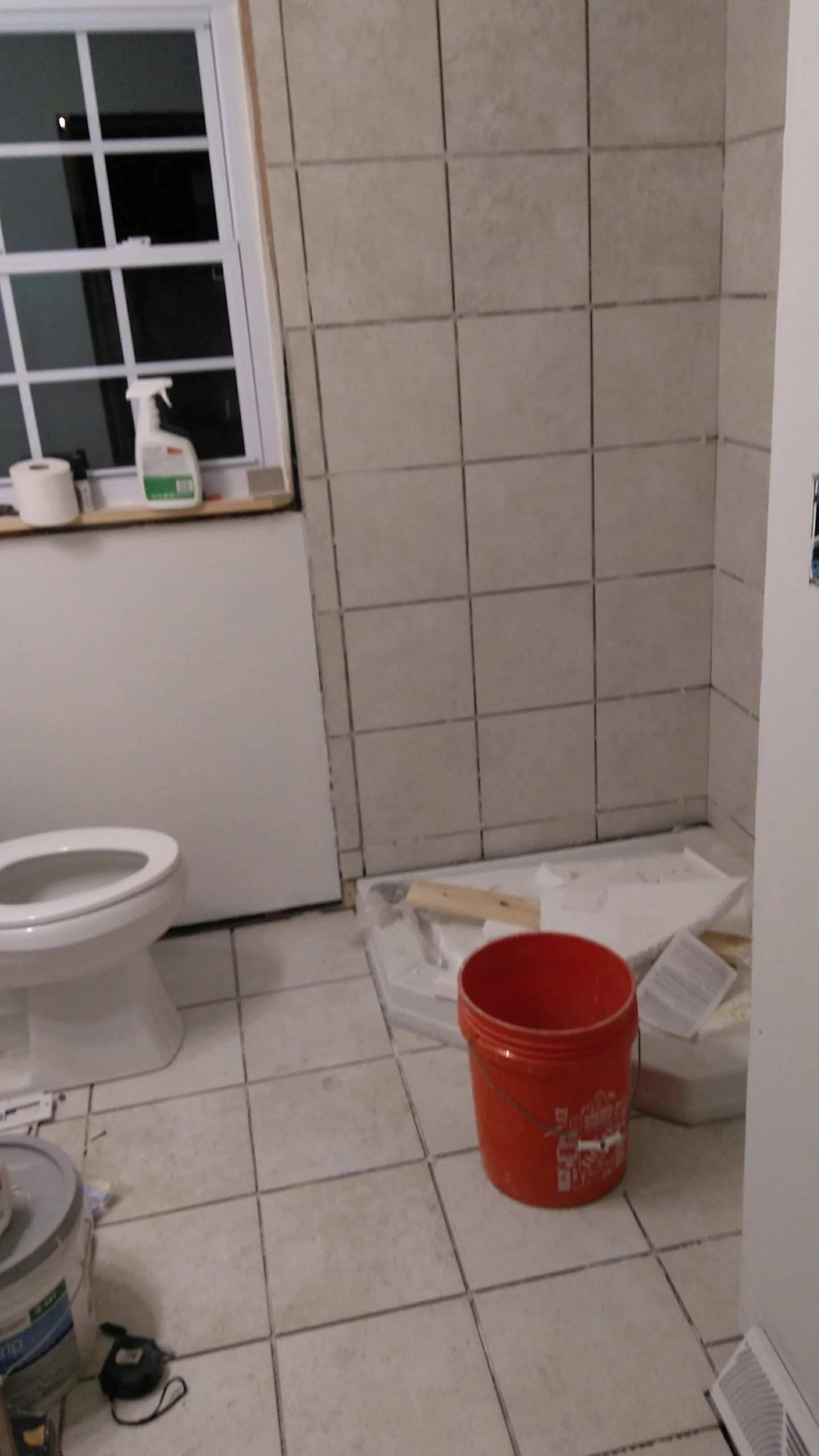 A bathroom under renovation with a toilet, construction supplies, a window, and a partially tiled shower area.
