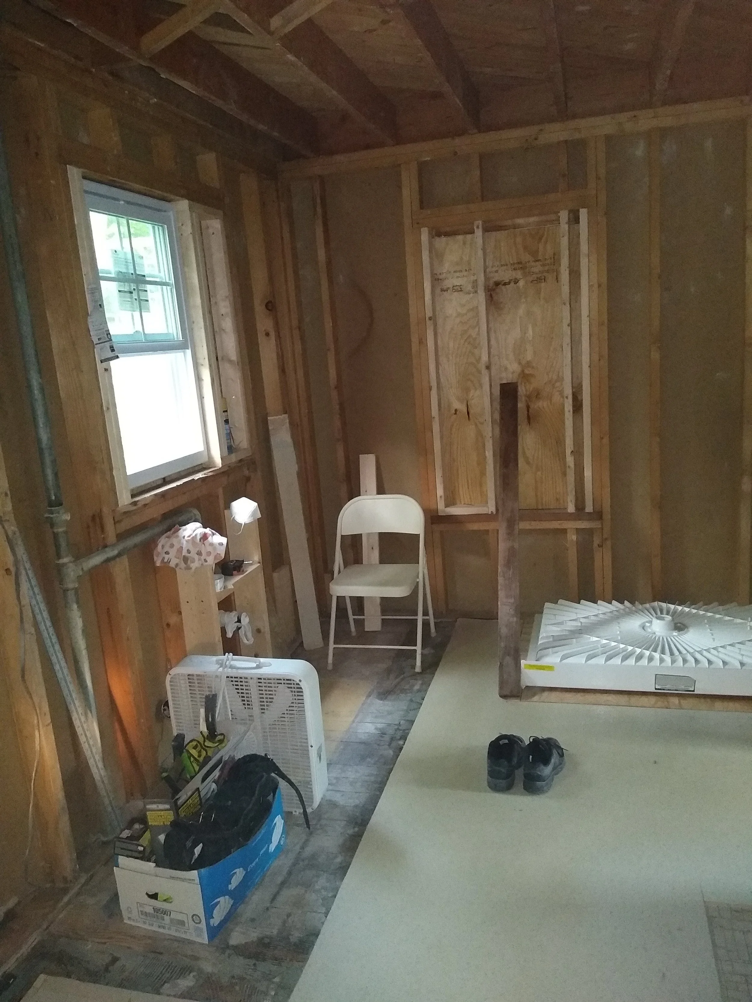 An unfinished room with exposed wooden framing and a small window. There is a white folding chair, a box with tools, a white fan, and a pair of black shoes on the floor. Construction materials and equipment are visible.