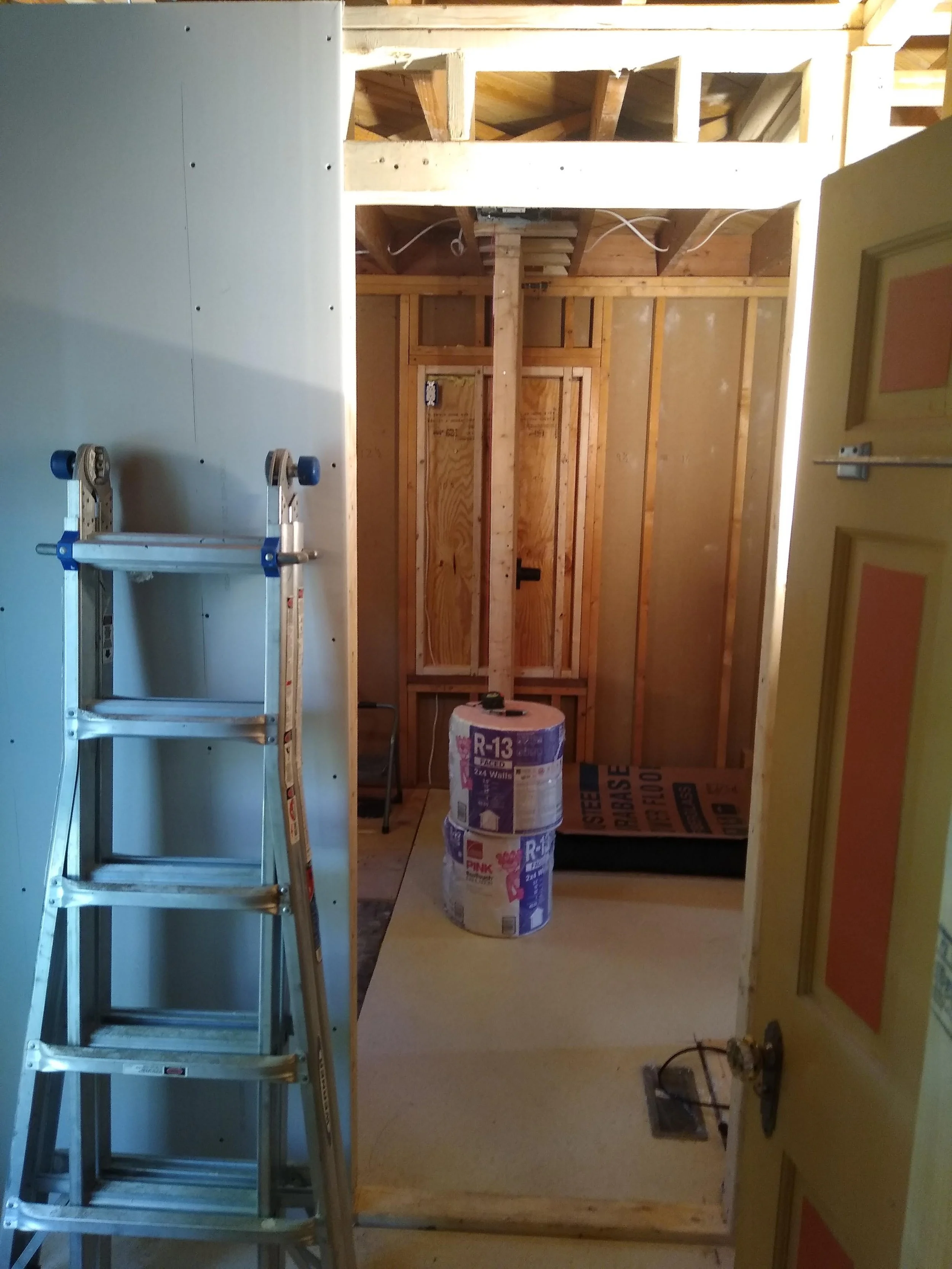 A room under construction with wooden framing, a stepladder on the left, and insulation rolls stacked in the center.