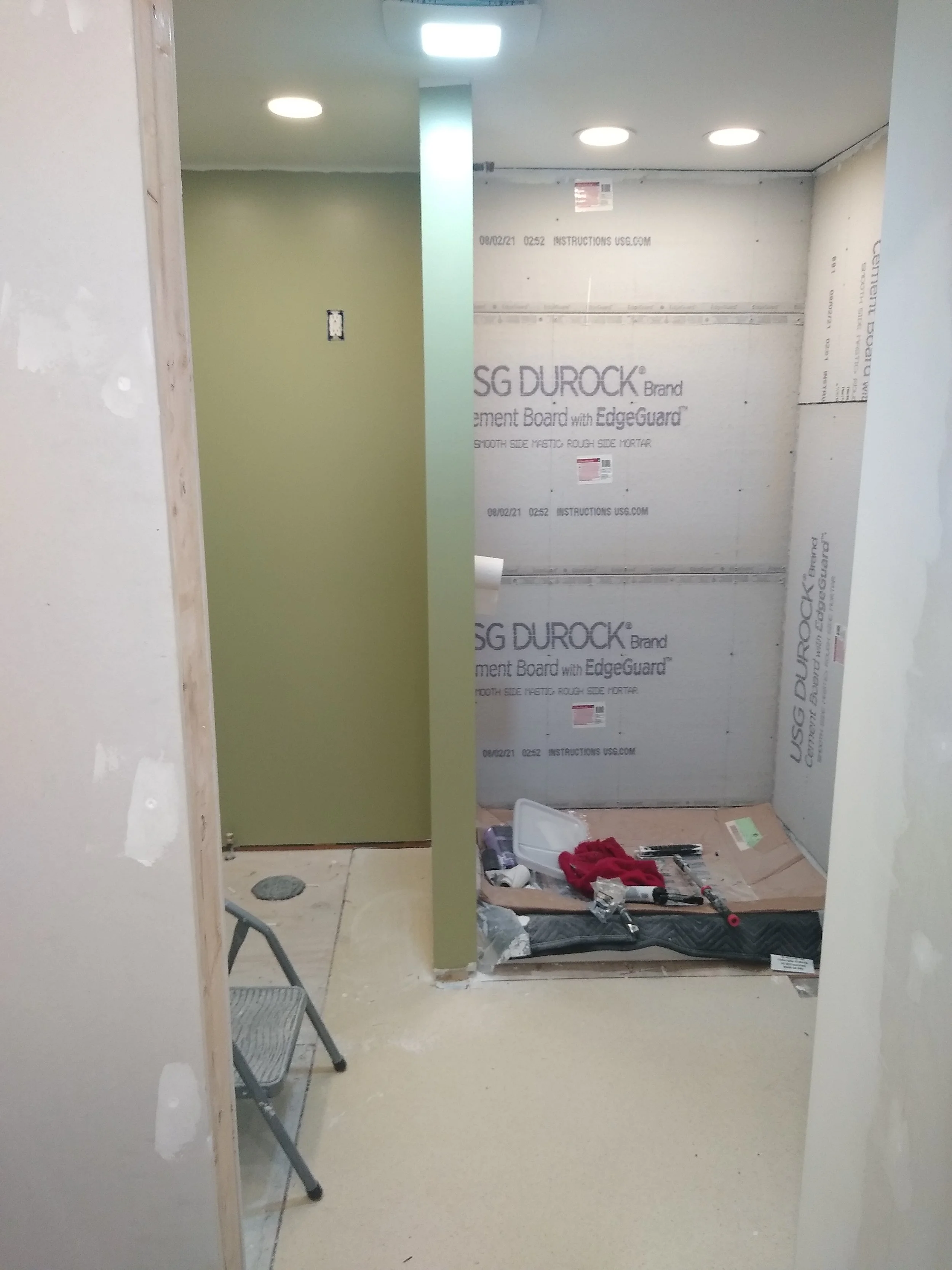 Construction site with partially installed green door and wall, drywall with protective covering, construction tools, and a small step ladder.