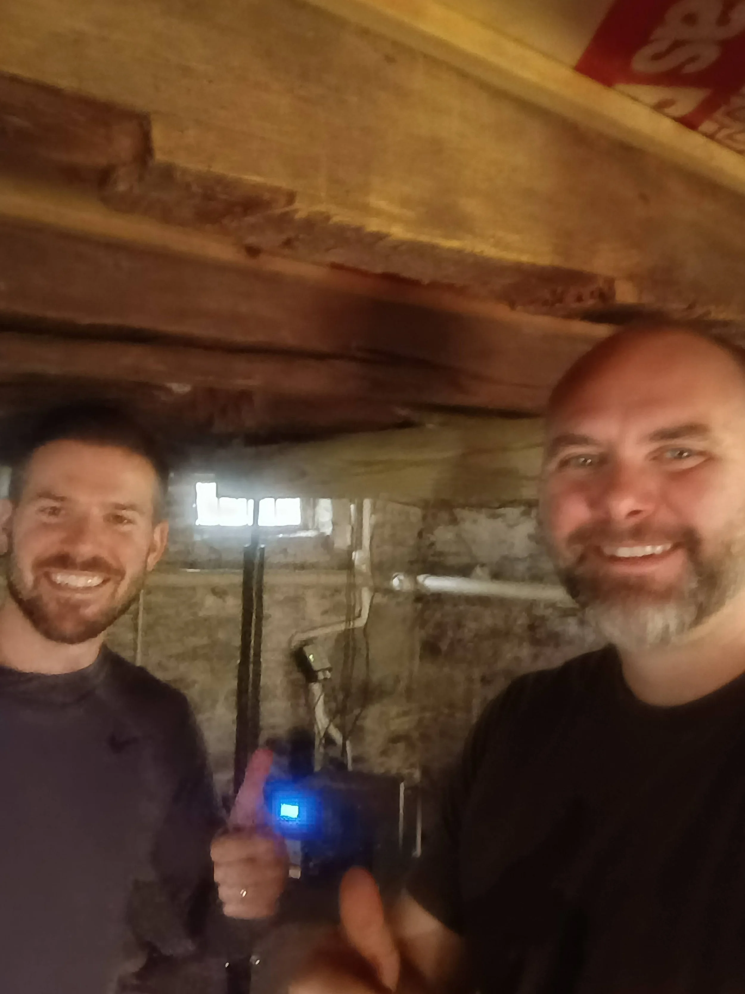 Steve and associate smiling inside a rustic wooden room, posing for a selfie, with one giving a thumbs-up and holding some electronic device with a blue light.