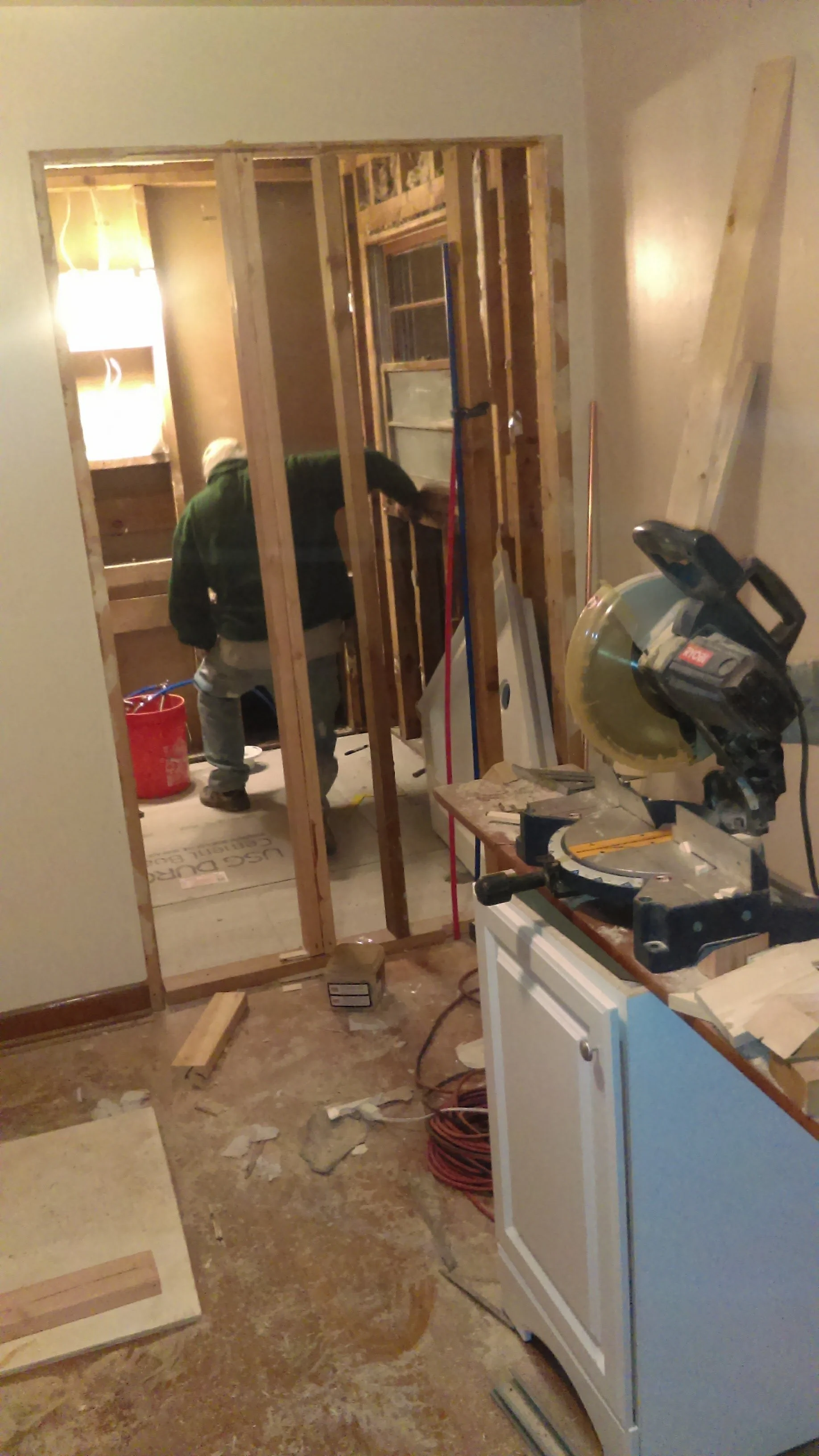 Interior view of a room under renovation with a person working behind a partially framed wall, surrounded by construction tools and materials.