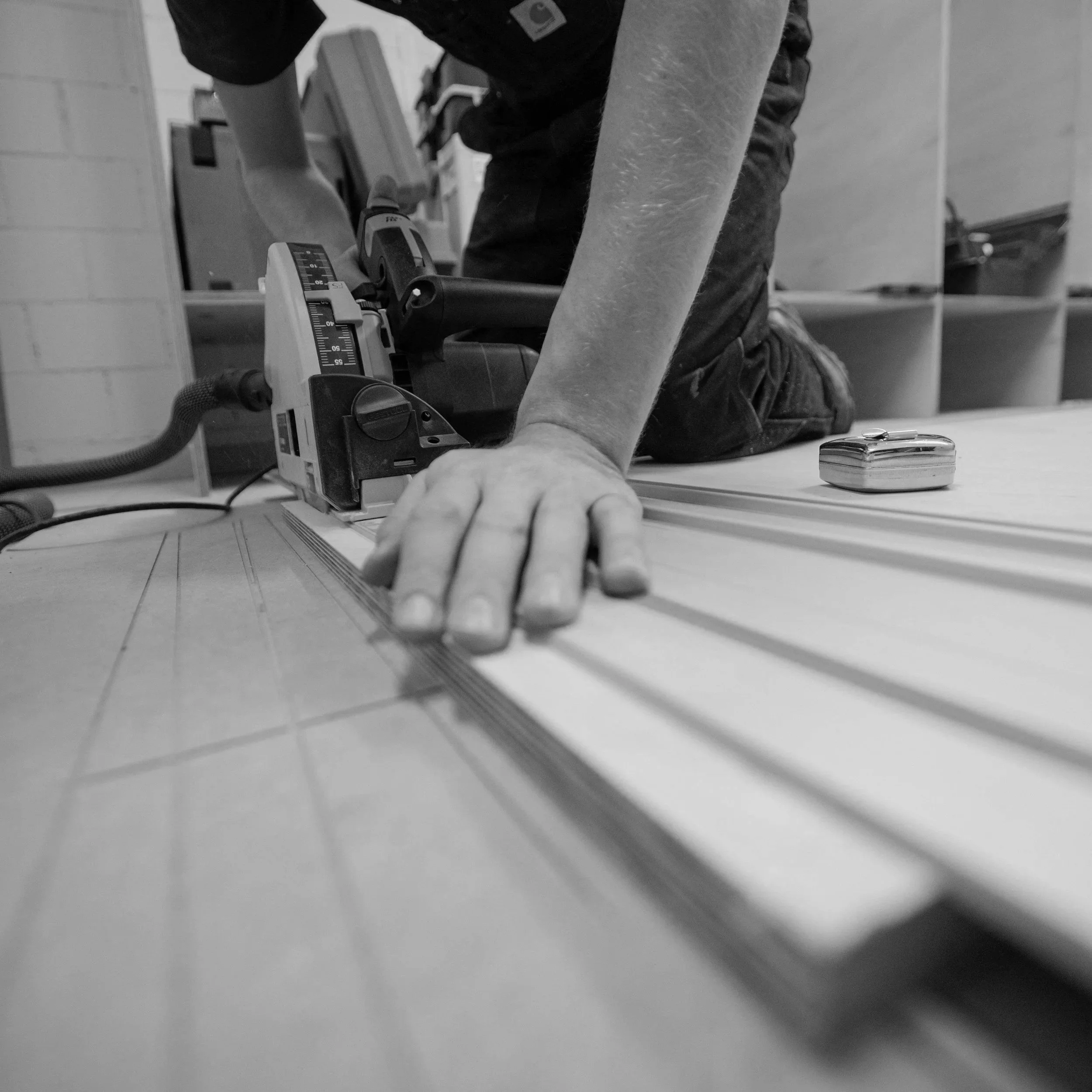 A person kneeling on the floor using a power tool to cut or shape a piece of wood with a measuring tape nearby in a workshop.