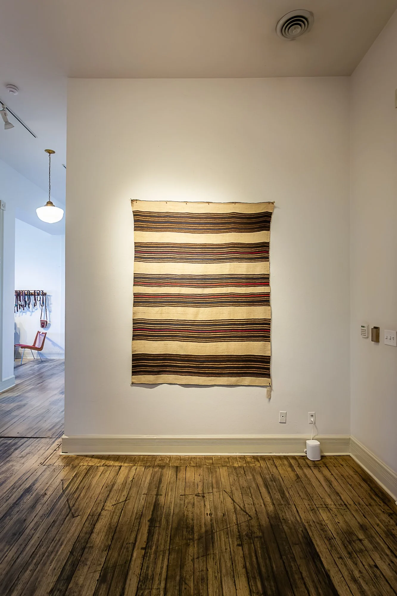 A striped Native American blanket Navajo Weaving wall hanging on a white wall in a room with wooden floors, with a small white device on the floor connected to a wall outlet.