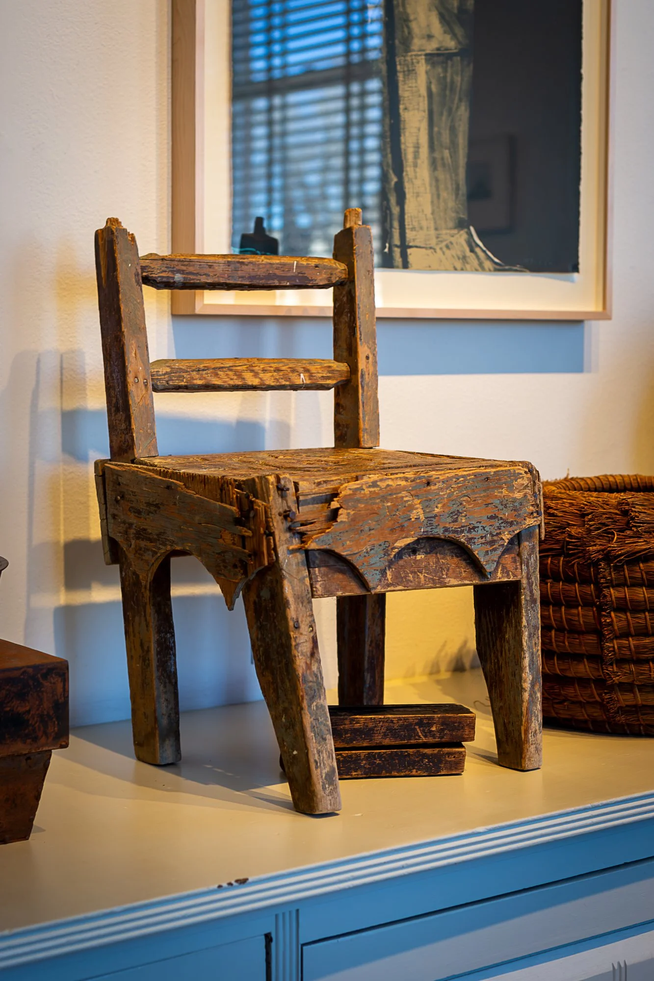 A small, rustic, wooden chair with a weathered and aged surface, placed on a white cabinet or sideboard, with a framed picture and a woven basket in the background.