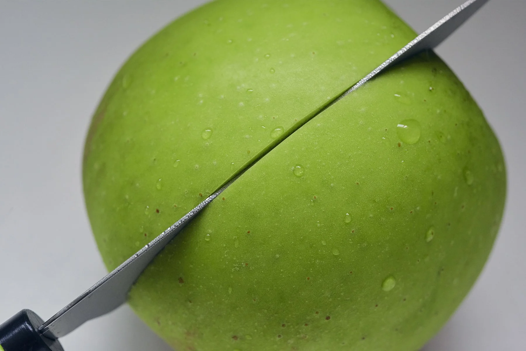 Close-up of a green apple being sliced with a knife, water droplets on the surface taken by Seoul based contemporary Korean Photographer Kim Sunik.