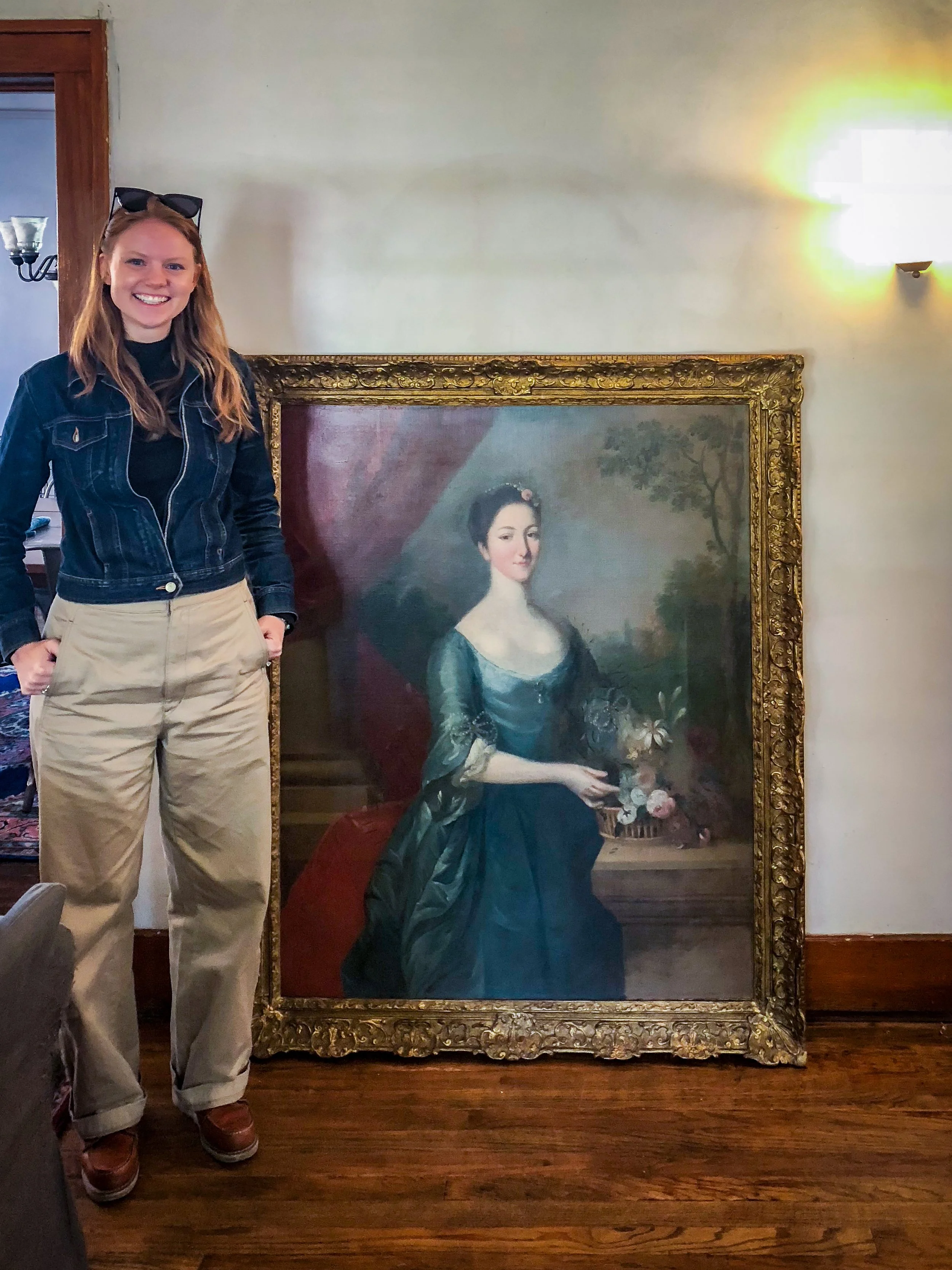 A smiling young woman standing next to a large portrait of a woman in historical attire in a framed painting, indoors with wooden flooring and a textured wall.