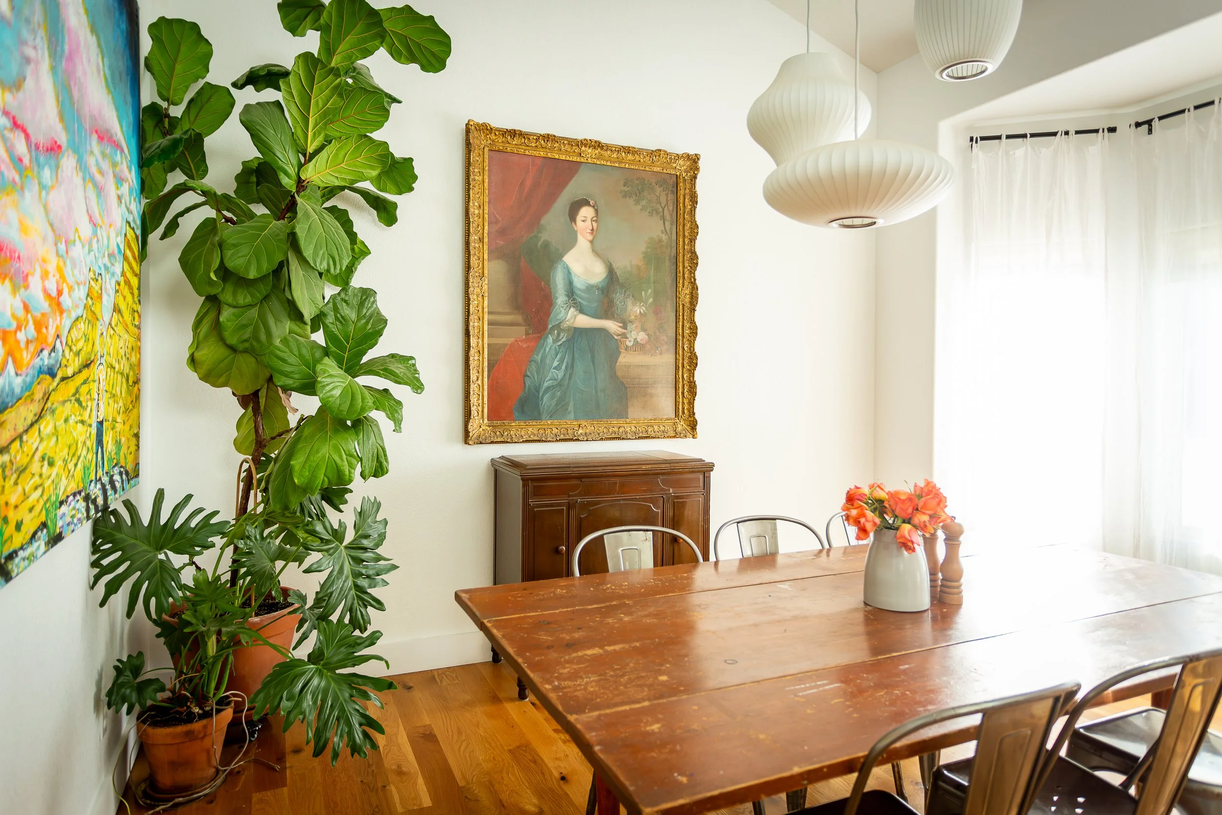 A cozy dining room with a large wooden table, Herman Miller Nelson pendant lights, a flower vase with pink flowers, a large leafy potted plant, a painting of a woman in a blue dress on the wall, once thought to be painted by Gainsborough.