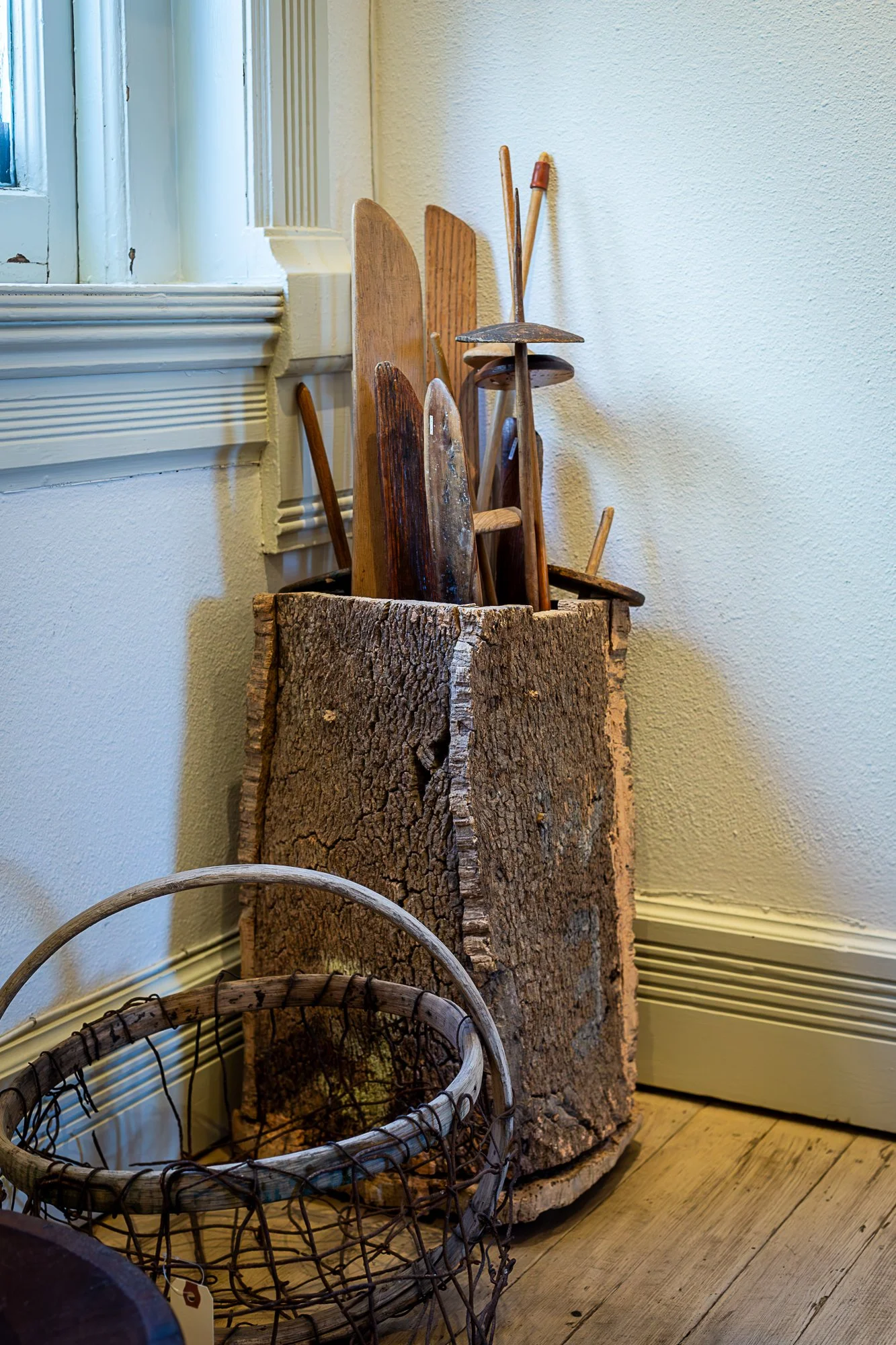 A wood log with various vintage wooden weaving battens and tools, next to a wire basket, placed inside a room with wooden flooring and a window.