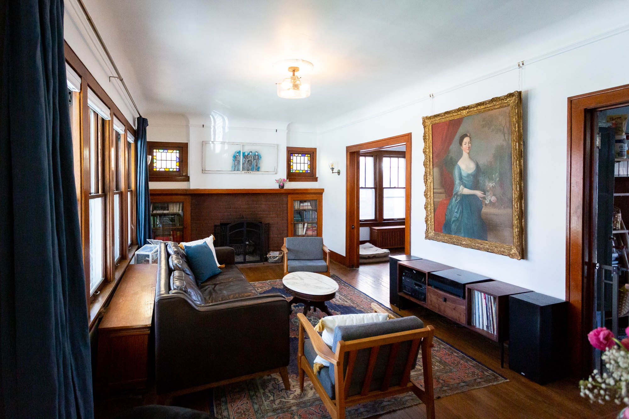 Craftsman style living room with large windows, dark leather sofa, armchair, fireplace, framed painting of a woman, wooden bookshelf, and stained glass windows in Detroit, Michigan.