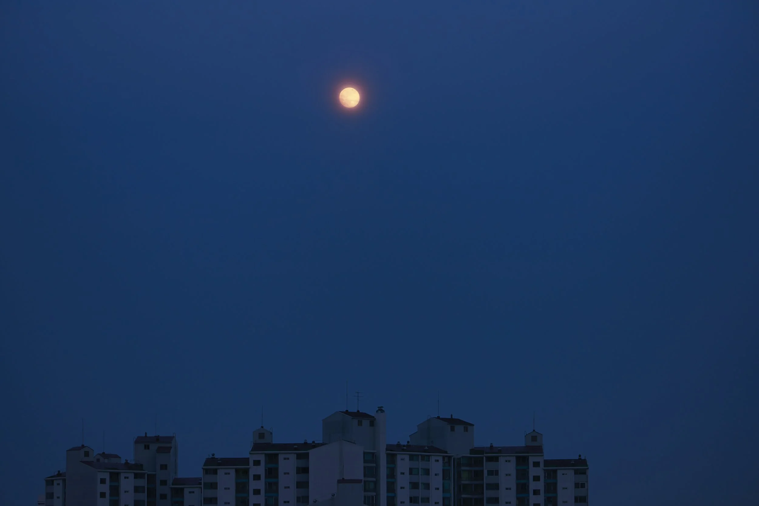 Nighttime scene with a bright full moon in the sky and a silhouette of a multi-story building below by Seoul based contemporary Korean Photographer Kim Sunik.