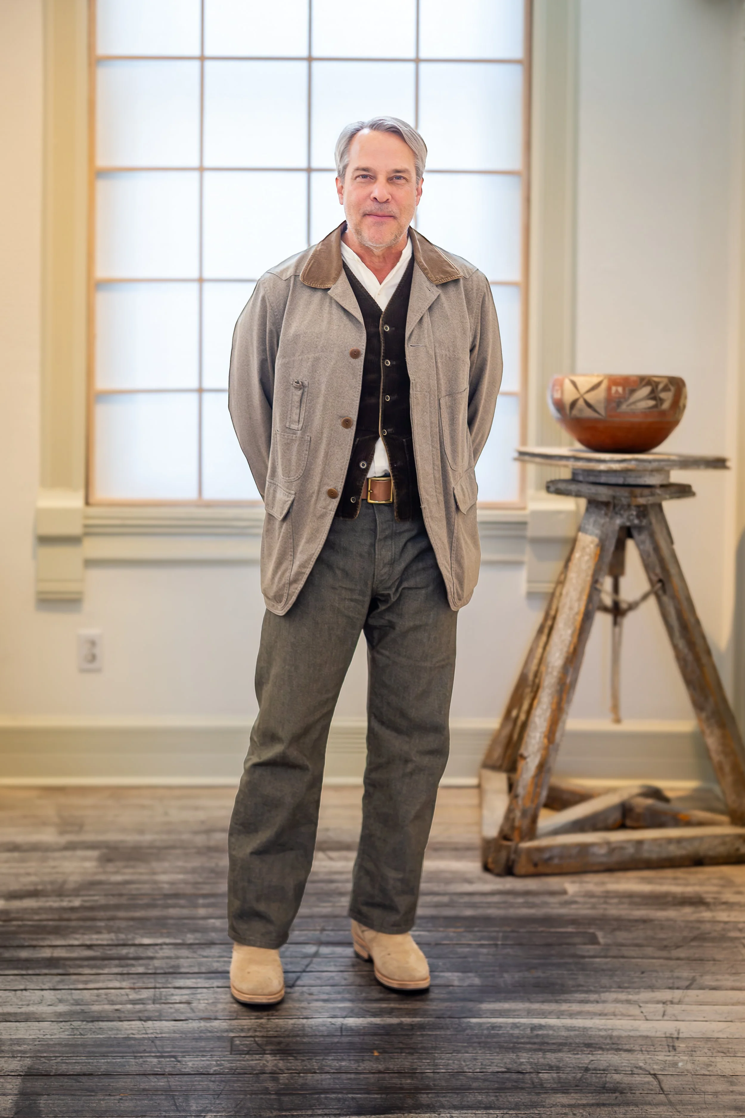 Jed Foutz, owner of Shiprock Santa Fe  stands in front of a window with frosted glass panes, wearing a beige jacket, black cardigan, white shirt, gray pants, and beige shoes in a well-lit room with wooden floors.