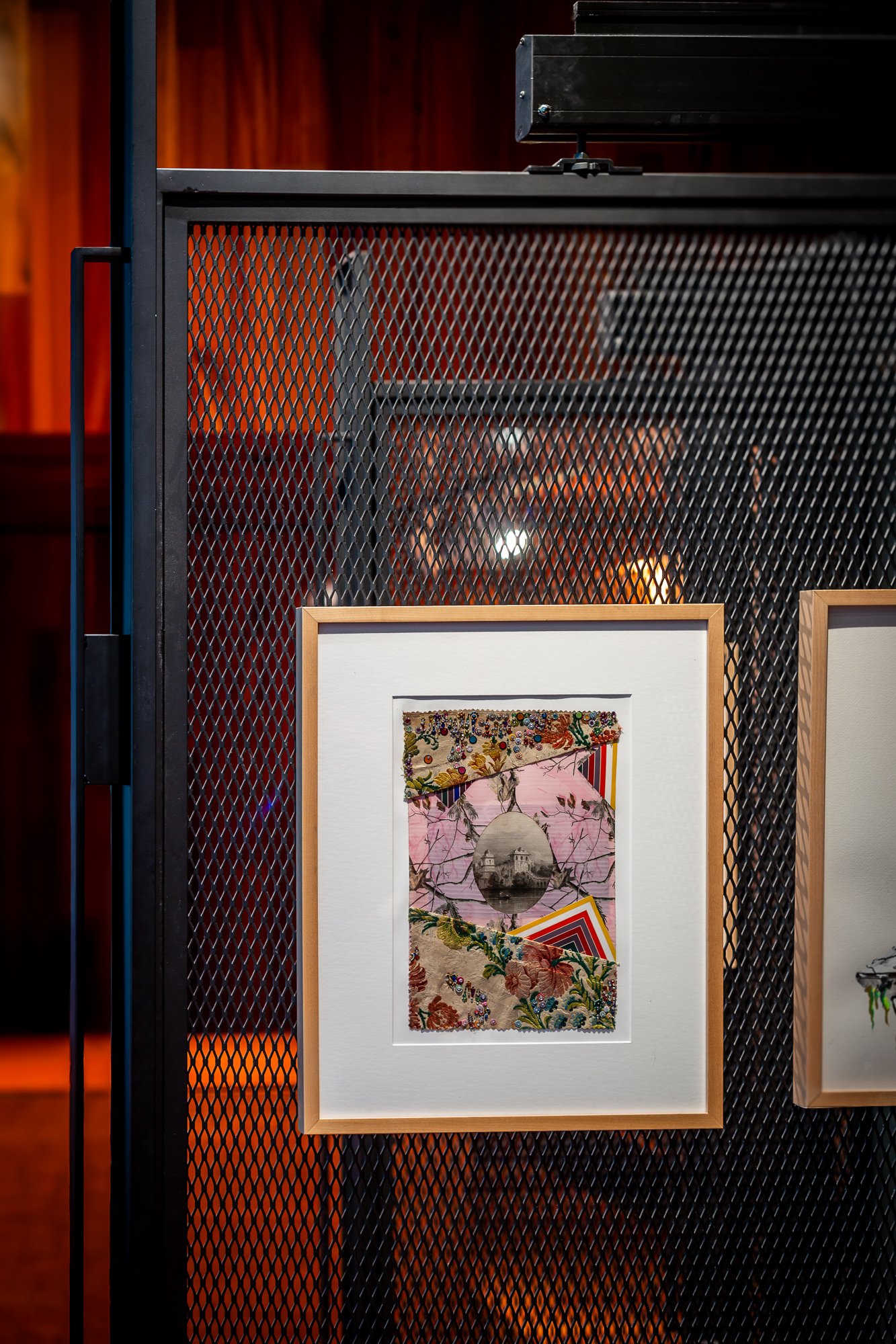 Artwork displayed on a black metal mesh stand with a wooden frame, featuring a collage with floral patterns, a vintage photograph, and geometric shapes, in an indoor exhibition setting.