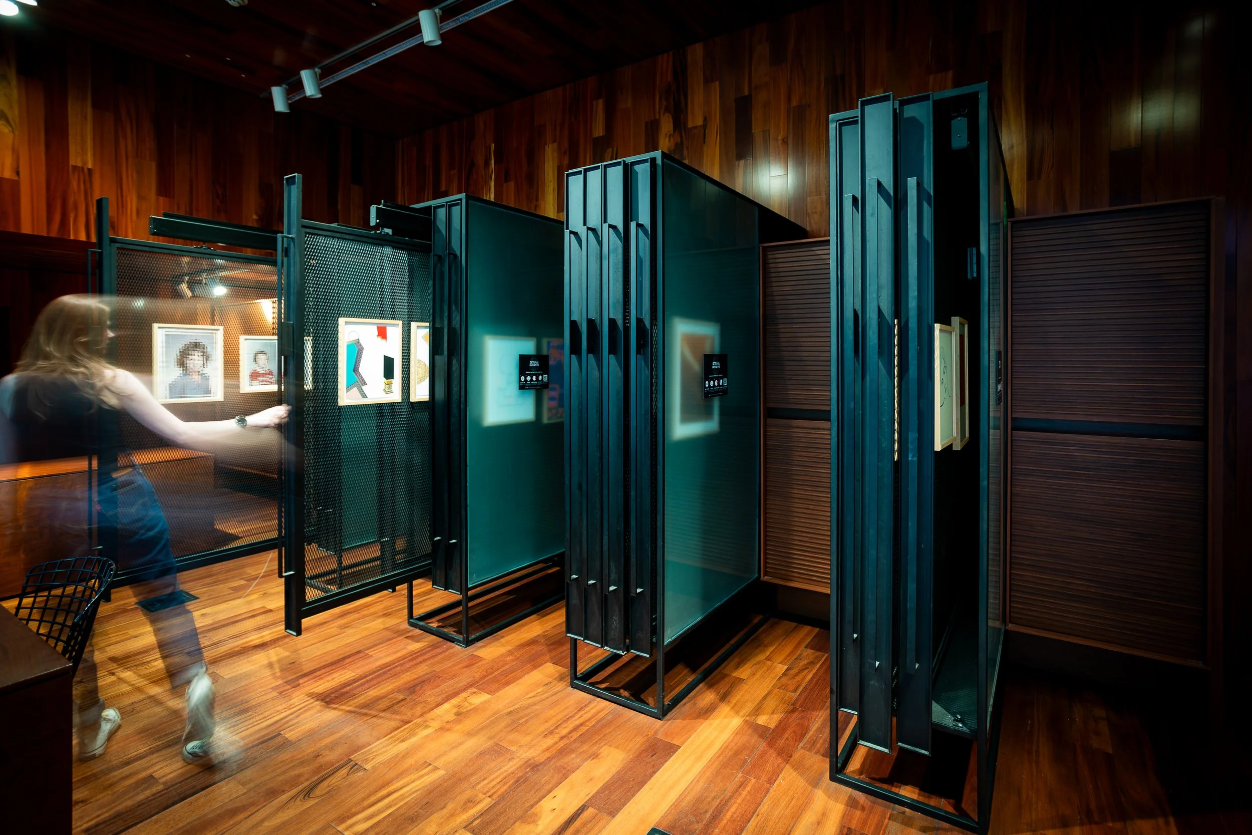 An art gallery exhibit featuring framed artwork displayed on black display boards, with a woman observing the art, in a room with wooden walls and floors.