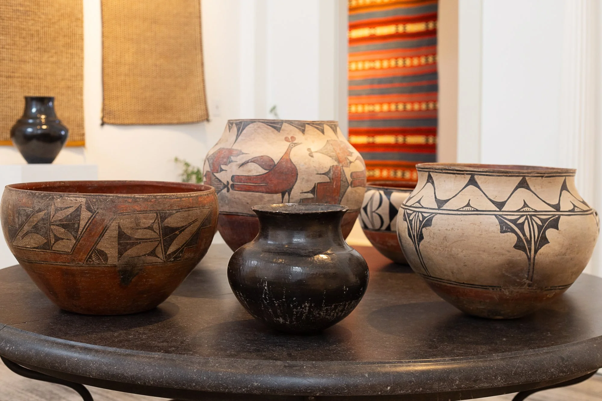 A collection of five antique native american zuni pottery vases with tribal patterns and designs on a dark circular table, with textiles and artworks in the background.