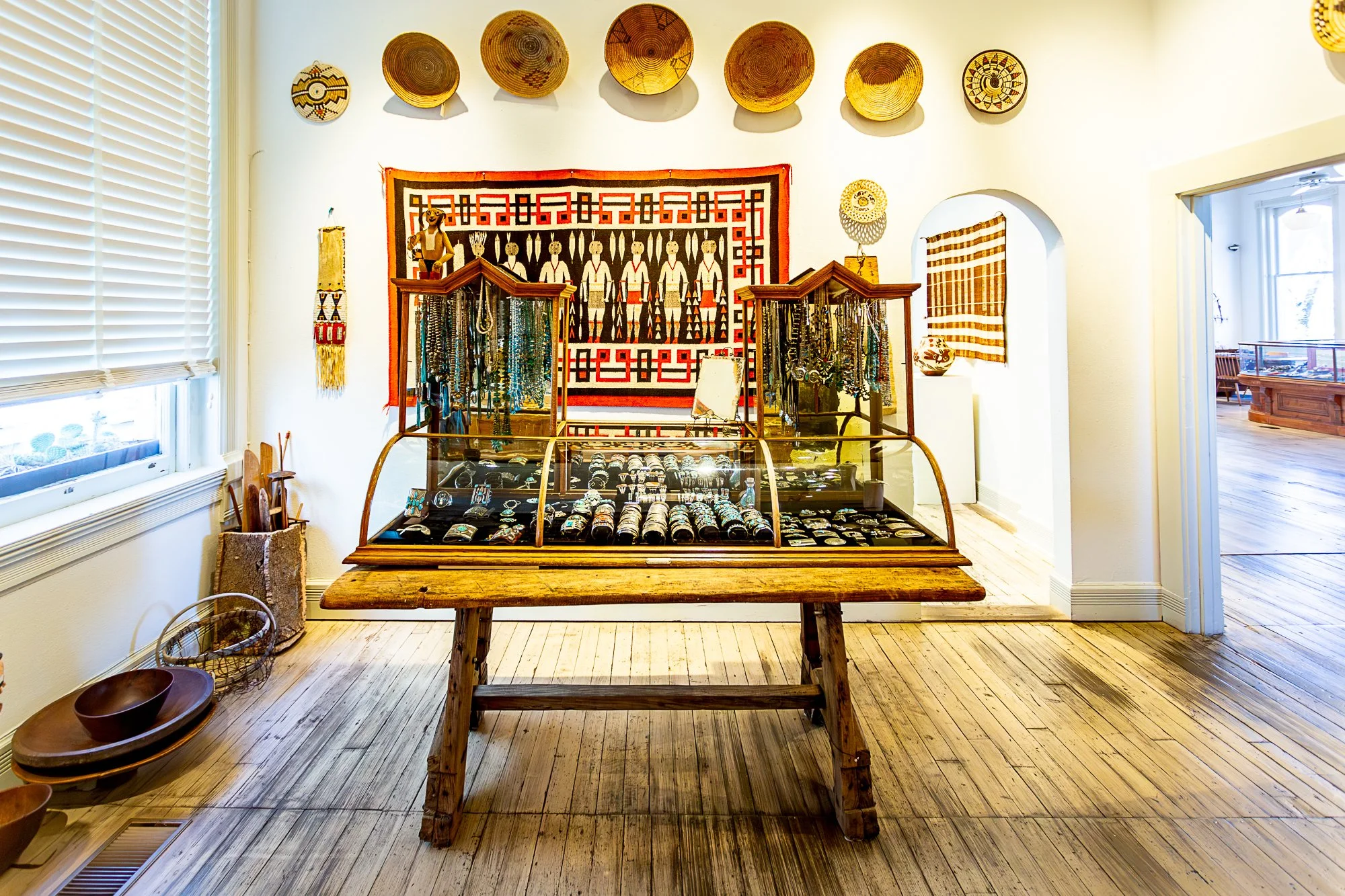 An interior view of a small shop displaying handmade jewelry and cultural artifacts. The shop features a glass display table with jewelry, woven baskets on the floor, and decorative wall hangings and baskets on the wall.