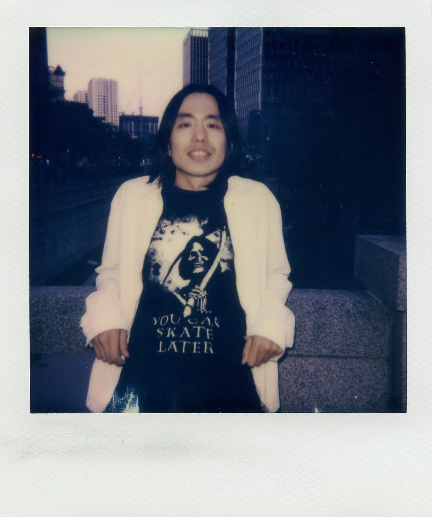 A person, Kim Sunik, standing outdoors in an urban area, wearing a black t-shirt with a graphic of a woman with a bandana and the text "You can skate later," a white jacket, and light-colored pants. The person has long dark hair and is smiling.