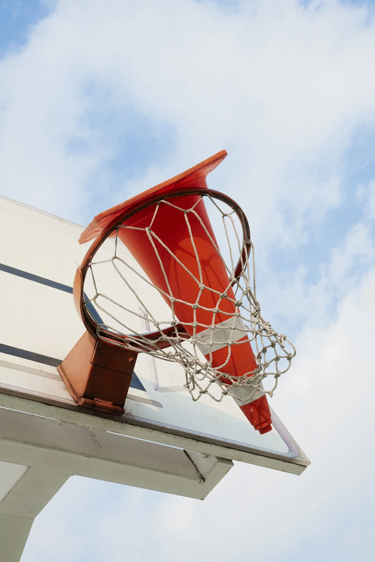 Low-angle shot of an outdoor basketball hoop with orange rim and net against a cloudy sky by Seoul based contemporary Korean Photographer Kim Sunik.