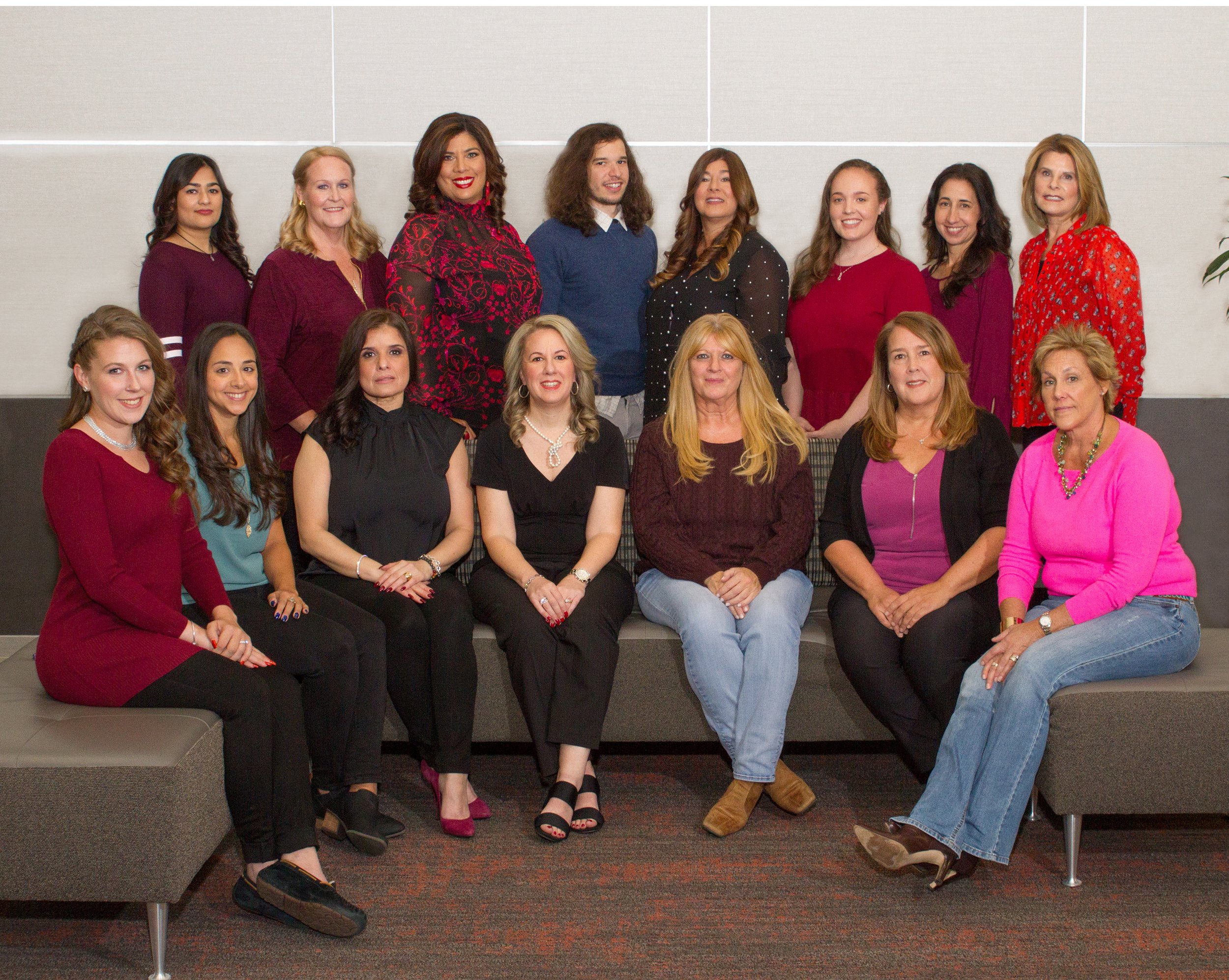 Group of 16 women and one man posing for a photo in an indoor setting, some sitting on a bench and others standing behind them, dressed in casual and business casual attire.