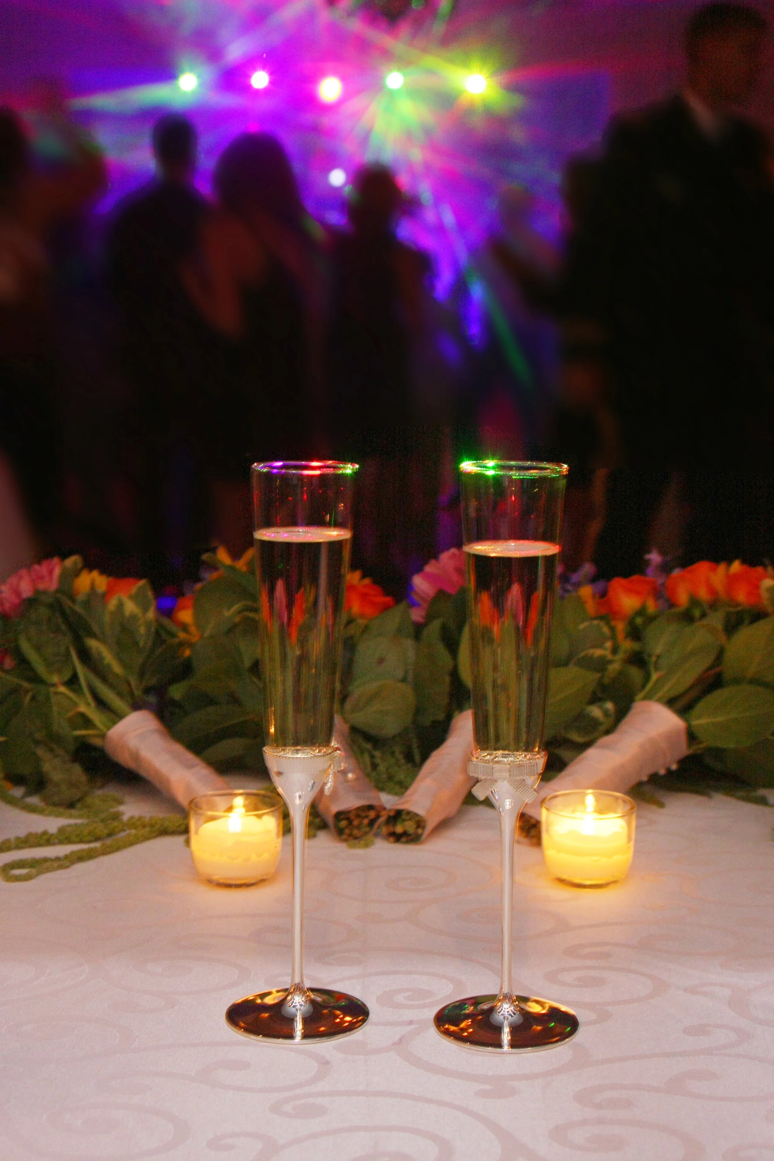 Two glasses of champagne on a decorated table with candles, a bouquet of flowers, and a crowd dancing under colorful lights in the background at the Ramsey Golf and Country Club.