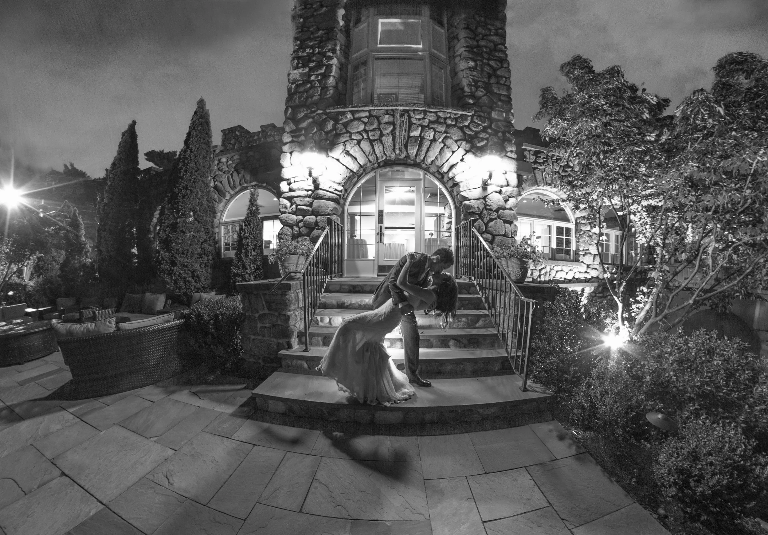 A couple is dancing on the steps outside a large stone house at night, illuminated by outdoor lights. The man is dipping the woman as they dance, with the woman wearing a long dress and the man in a suit. There is outdoor furniture and a tree decorat