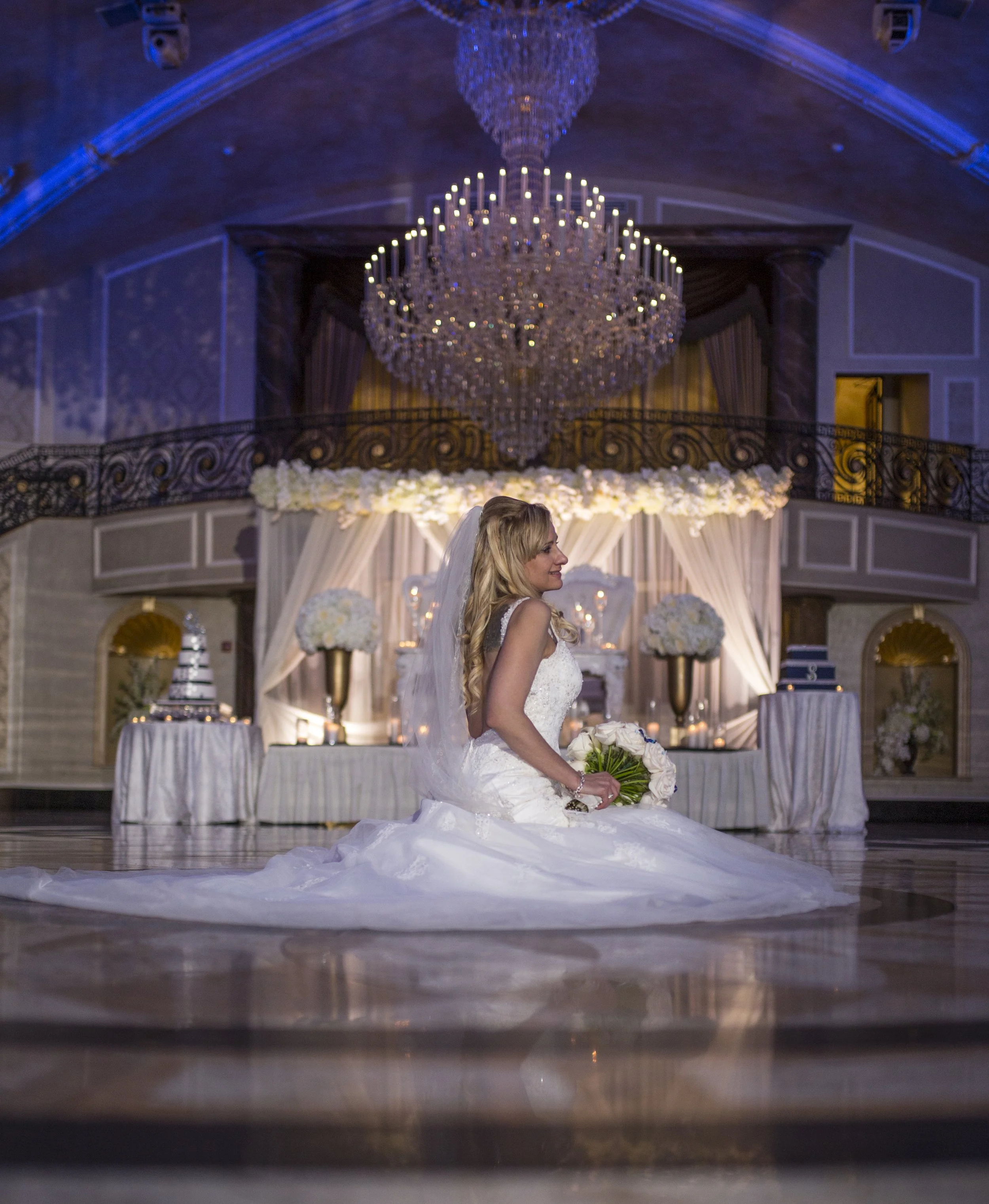 Bride kneeling on the wedding dance floor in a white wedding gown holding a bouquet of white flowers, with an elaborately decorated wedding reception hall in the background featuring a chandelier and floral arrangements.