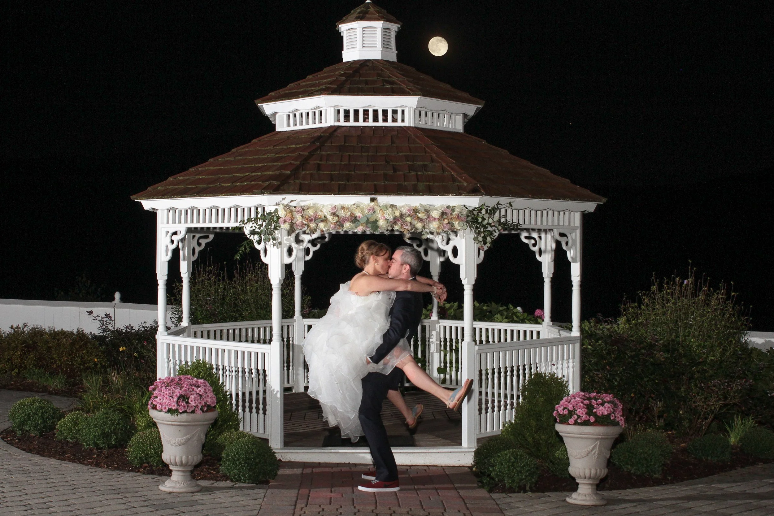 A man in a suit carrying a woman in a wedding dress inside a white gazebo at night with a full moon in the background.