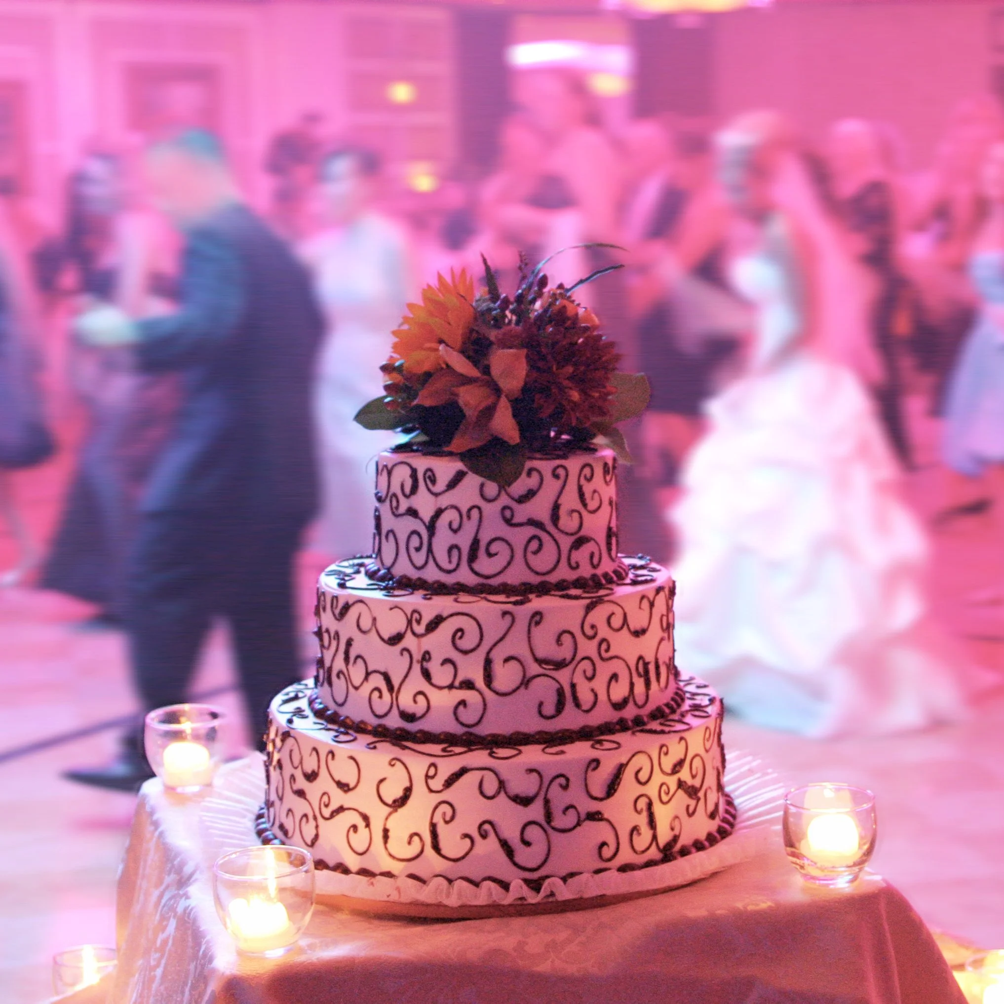 Three-tier wedding cake decorated with black swirl patterns, topped with a floral arrangement, on a table with small lit candles, and wedding guests dancing in the background.