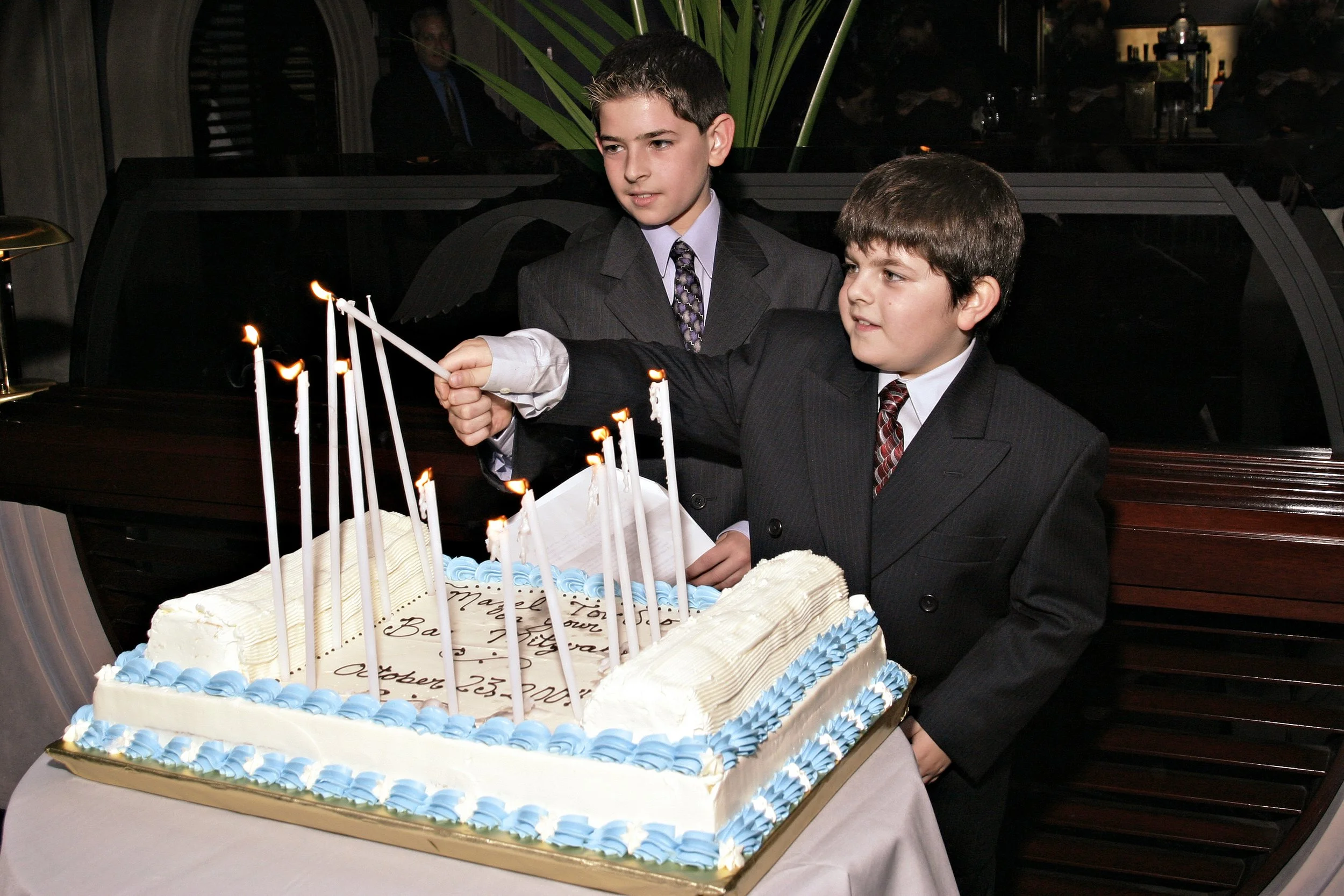Two boys in suits celebrating a Bar Mitzvah with a large decorated cake with lit candles.